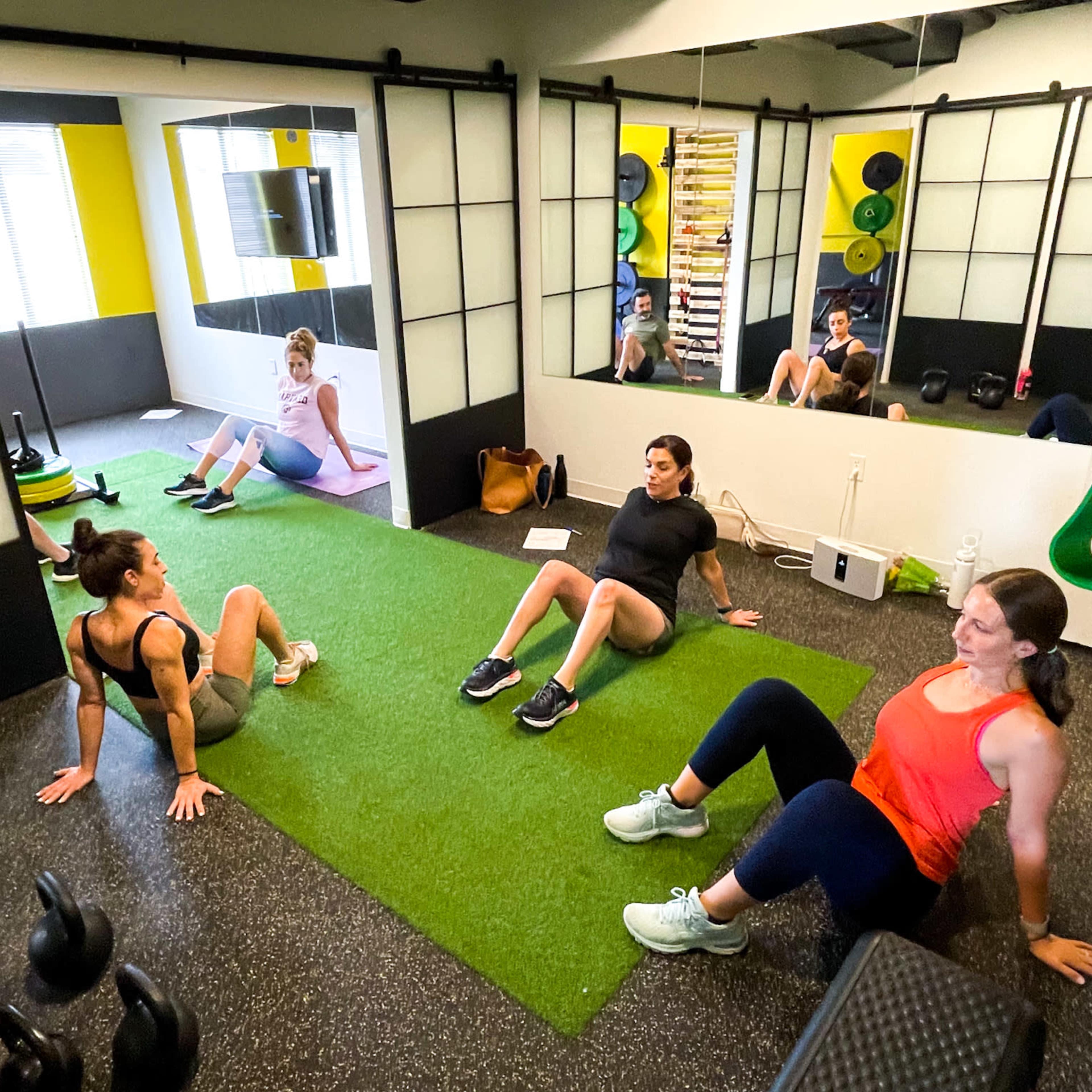 Three women are seated on a green exercise mat in a gym, while a fourth woman exercises in the background.