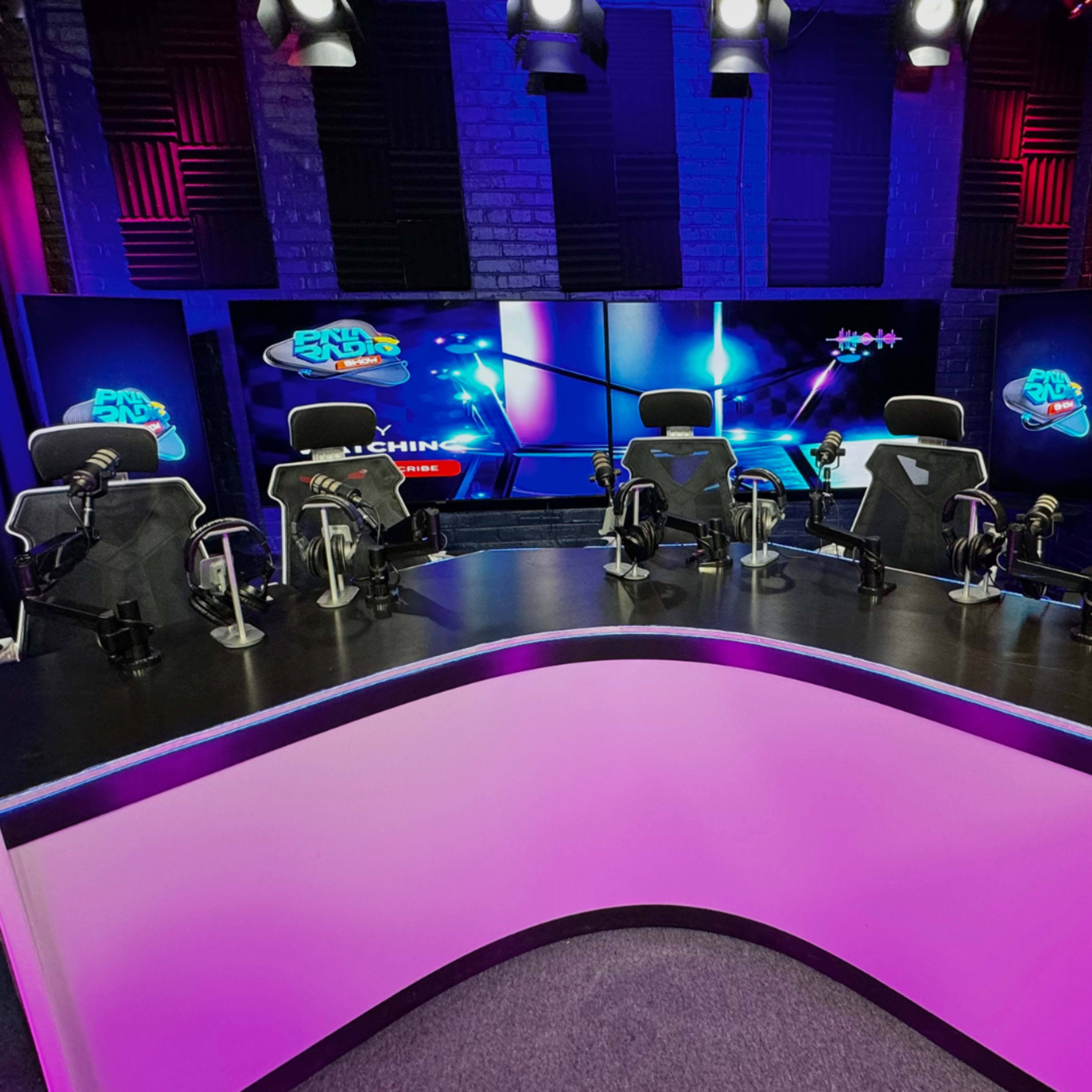 A modern studio setup with a central curved desk and several empty gaming chairs, illuminated by colorful lights and featuring large screens in the background.