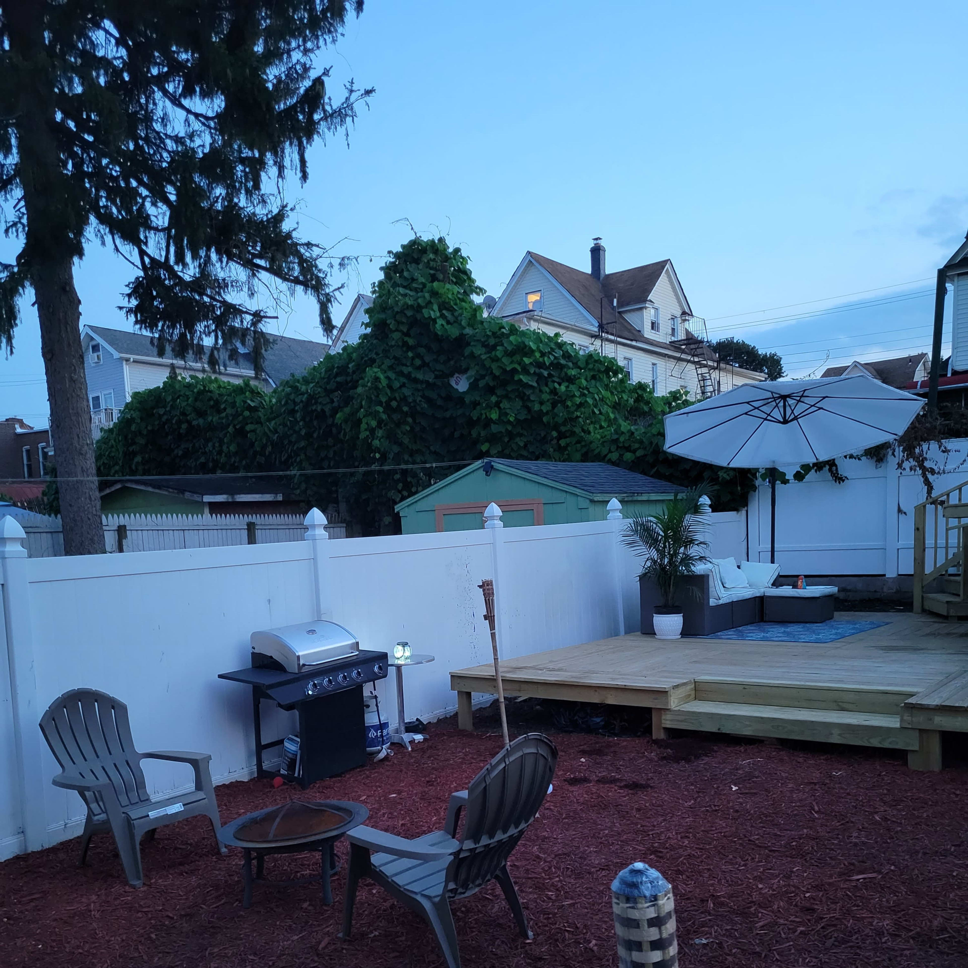 A backyard area with a grill, two chairs around a small fire pit, and a deck with an umbrella and a nearby storage shed.