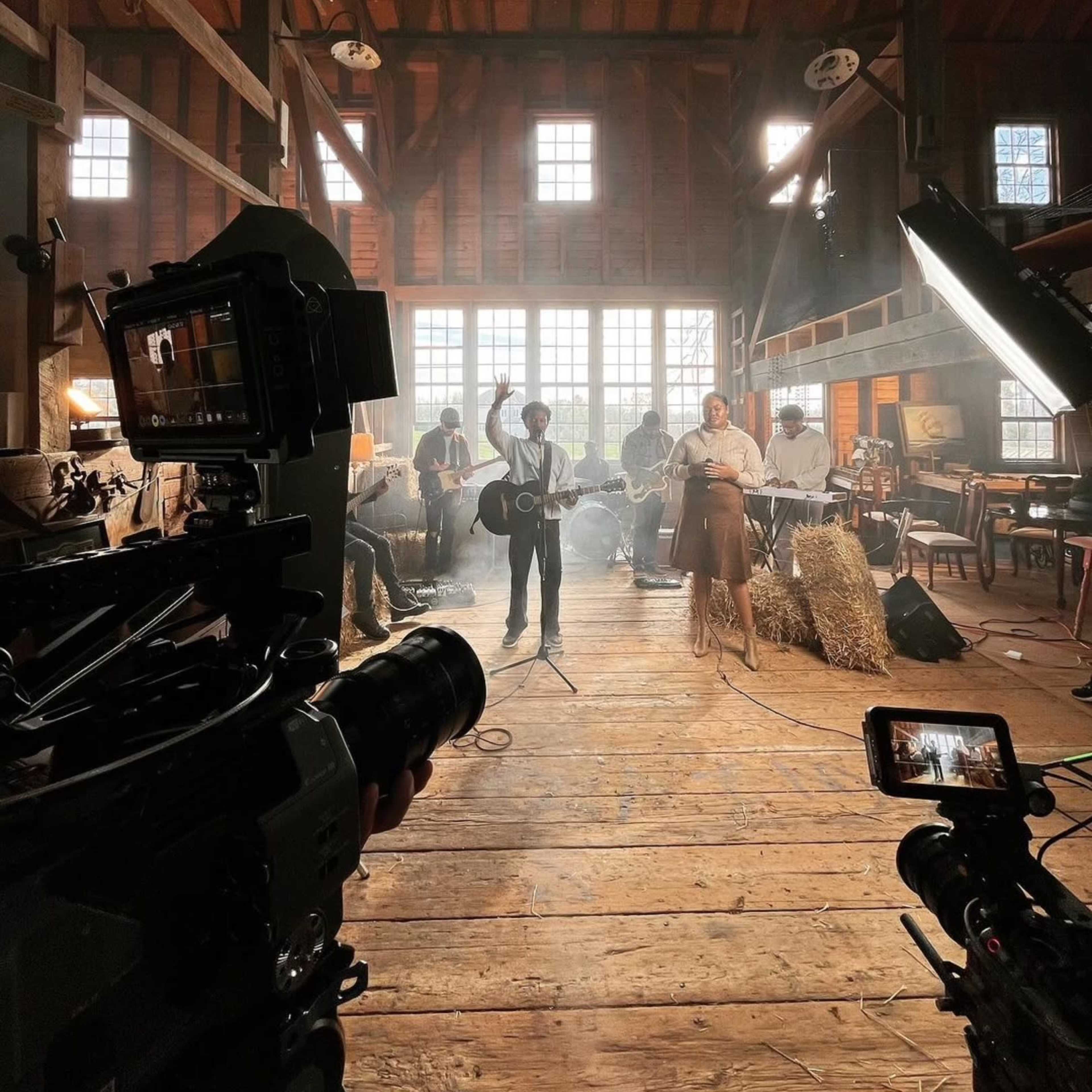 A musician stands in a barn-like space with a band, surrounded by cameras and lights.