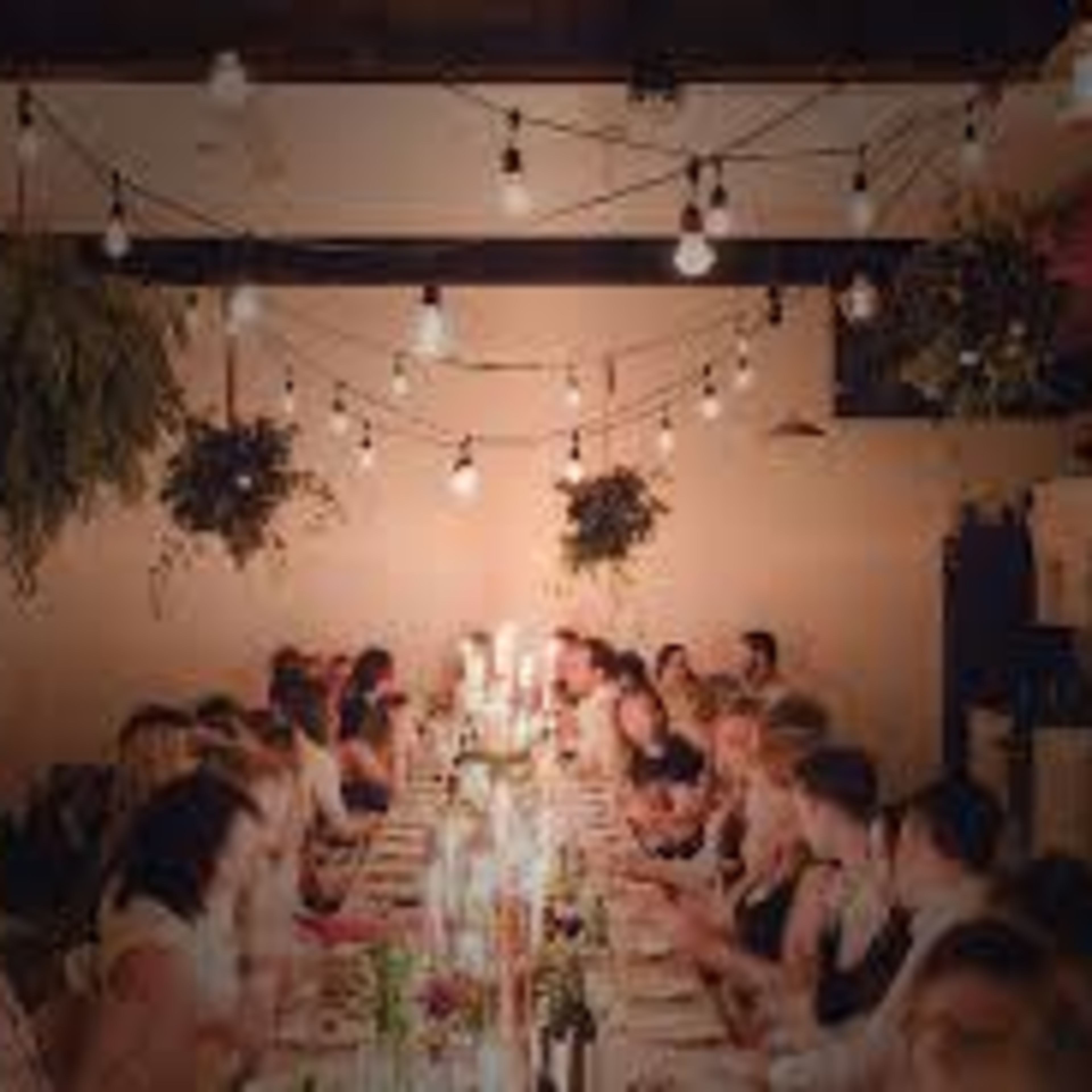 A long table is set for a dinner party, lined with candles and surrounded by guests in a warmly lit space with hanging plants and string lights overhead.