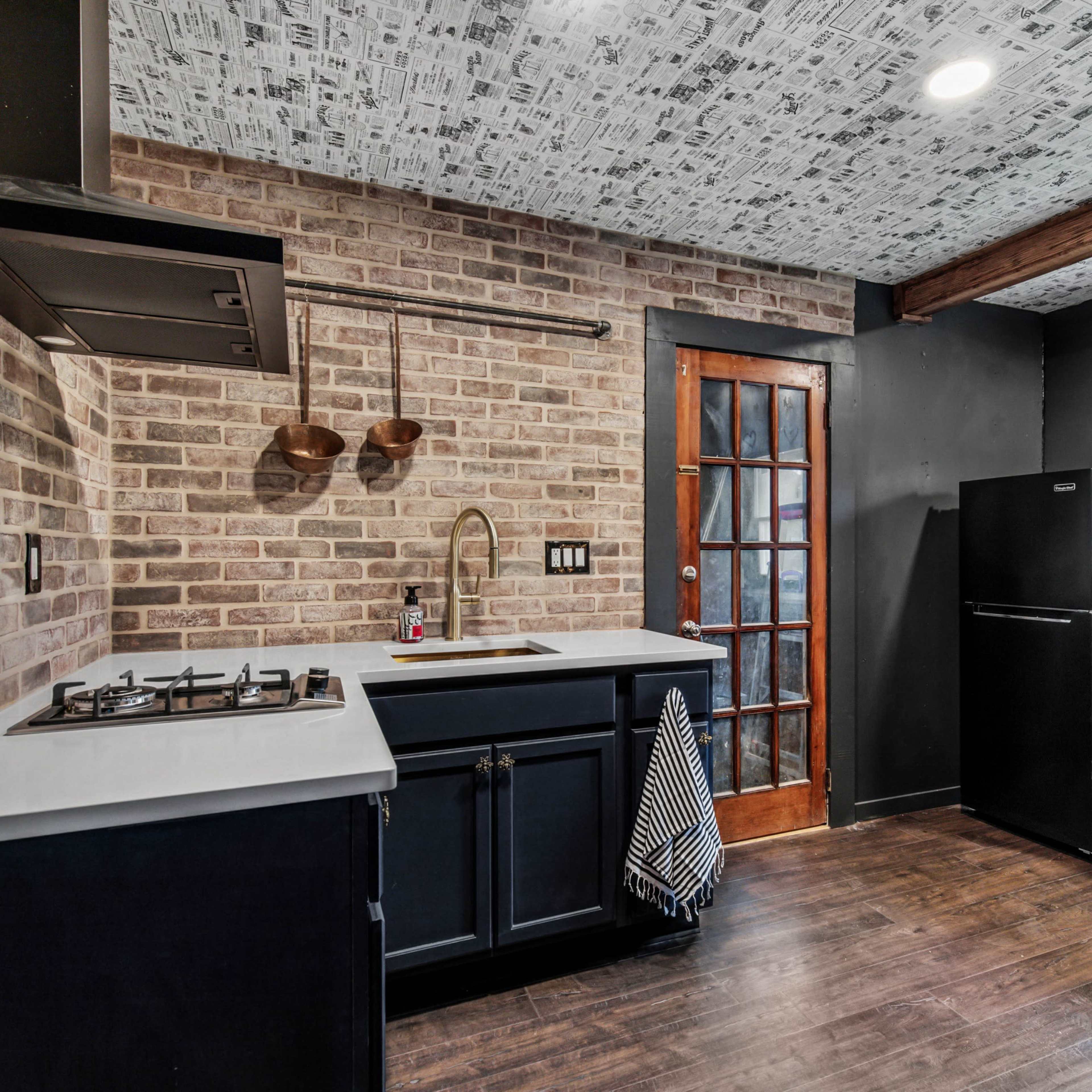 A modern kitchen with exposed brick walls, dark cabinetry, and a gas stove, featuring a wooden door and a black refrigerator.