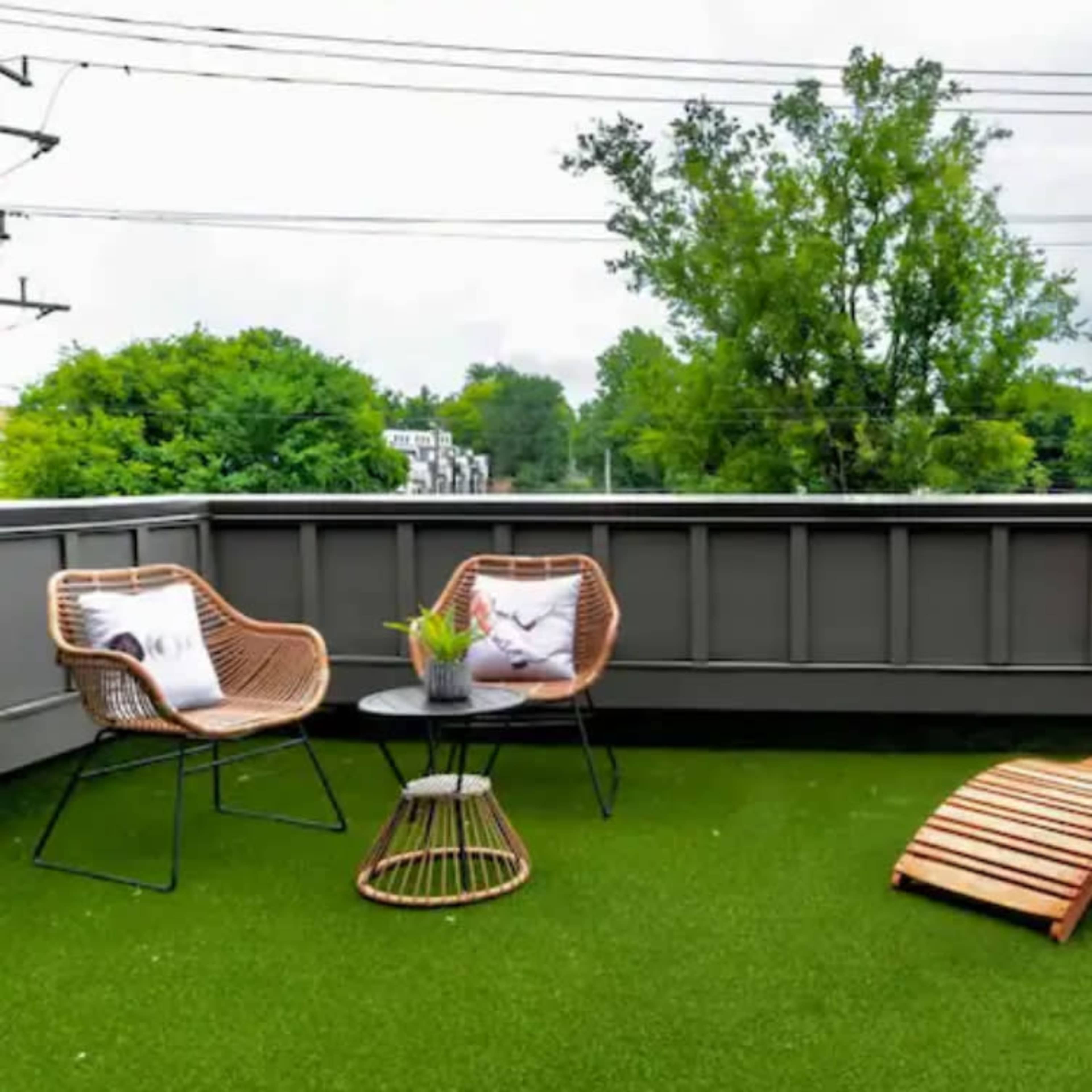 A rooftop patio features two lounge chairs, a small table, and decorative plants set on green artificial grass.