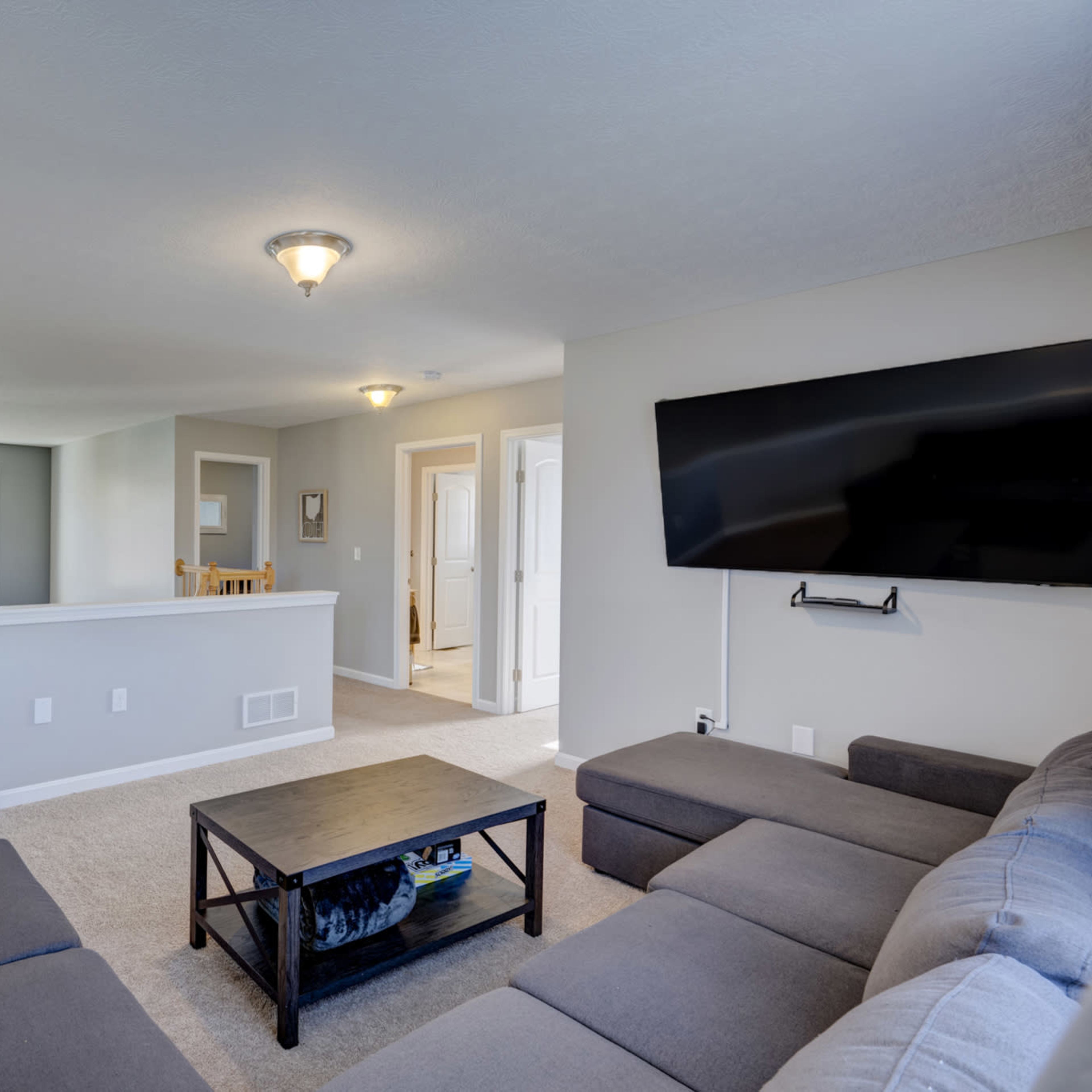 A modern living room with a sectional couch, a coffee table, and a wall-mounted television, connected to an open space with light-colored walls and windows.