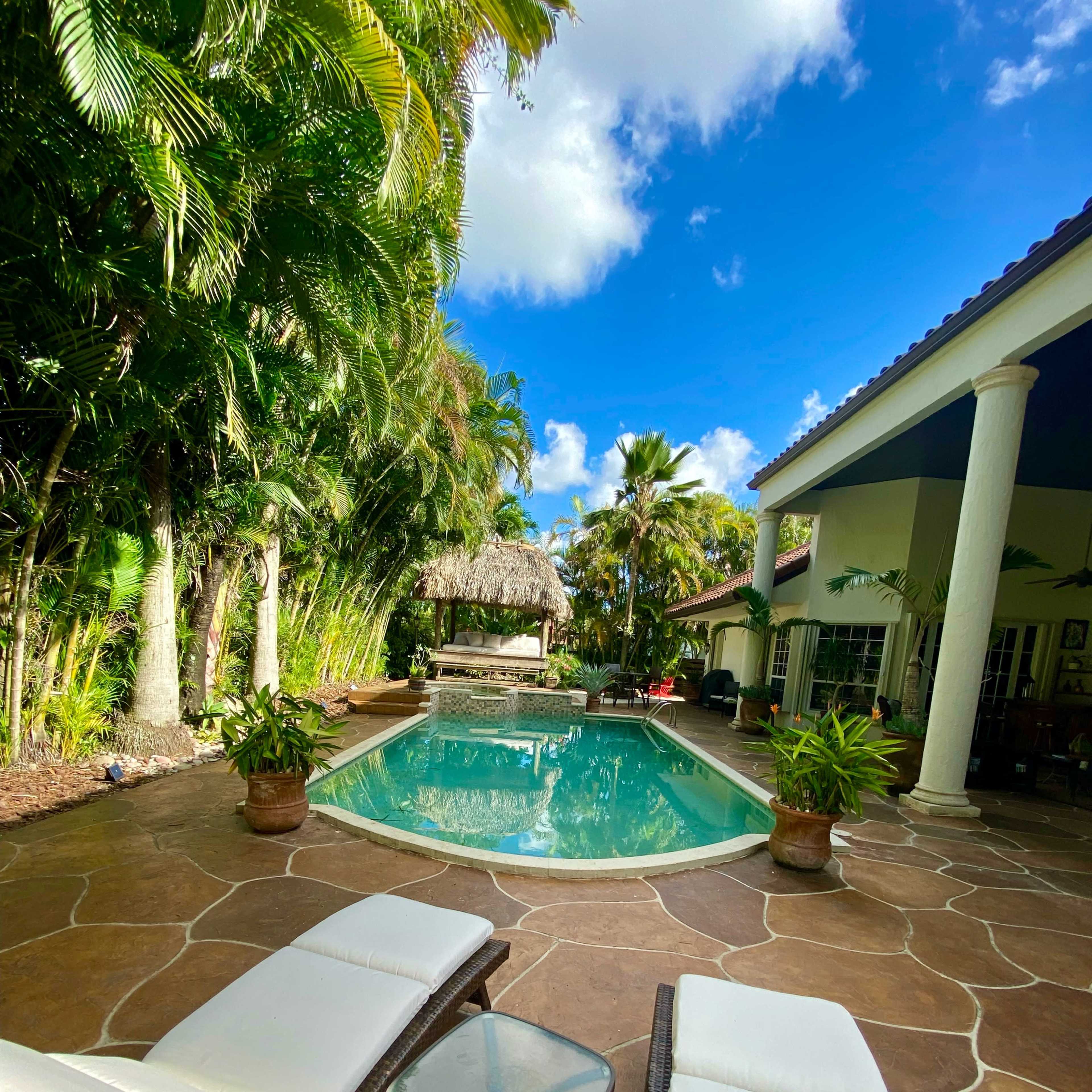 A pool surrounded by tropical plants and lounge chairs is located next to a house with a thatched roof area nearby.