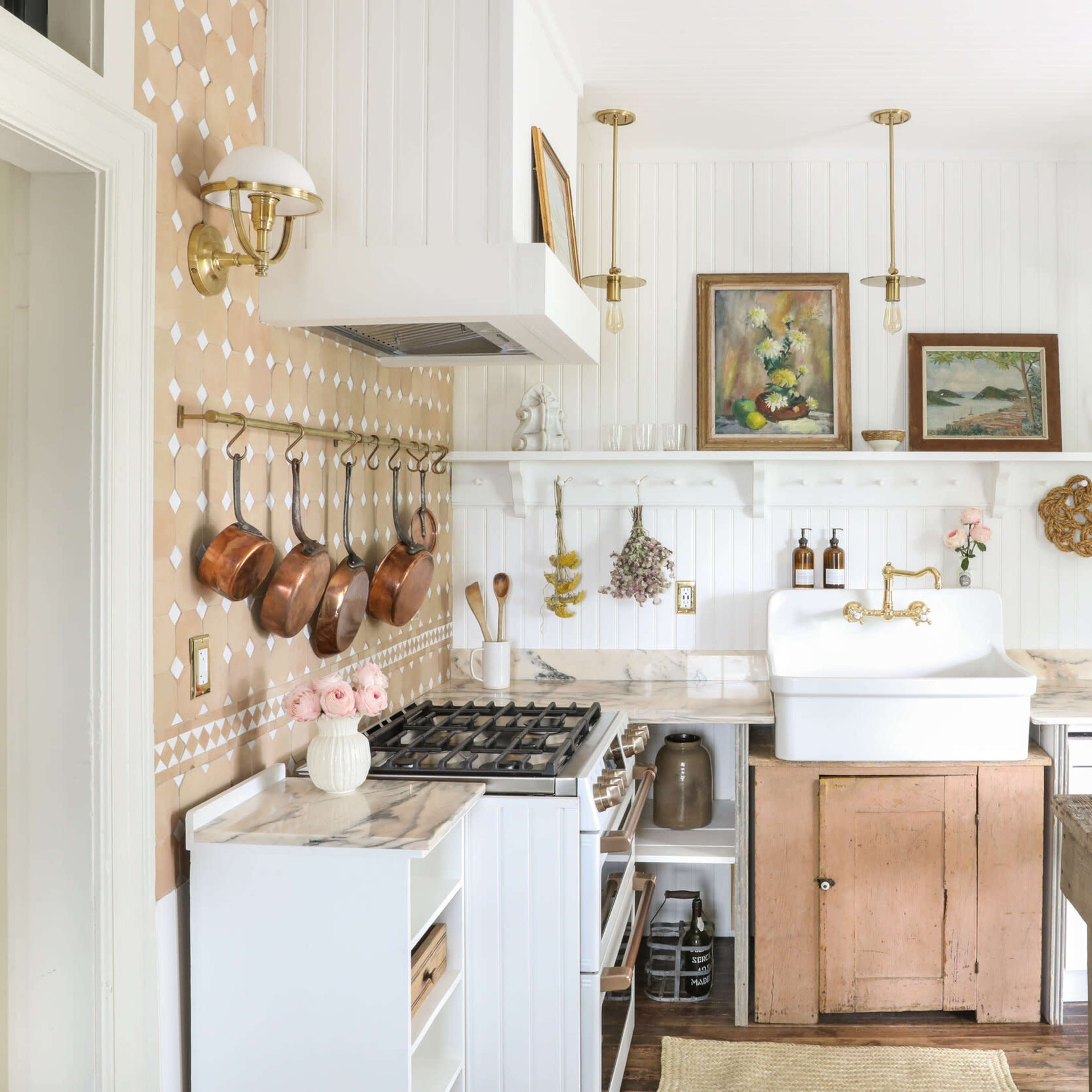 A bright kitchen features white walls, marble countertops, a farmhouse sink, and wooden accents with hanging pots and decorative artwork.