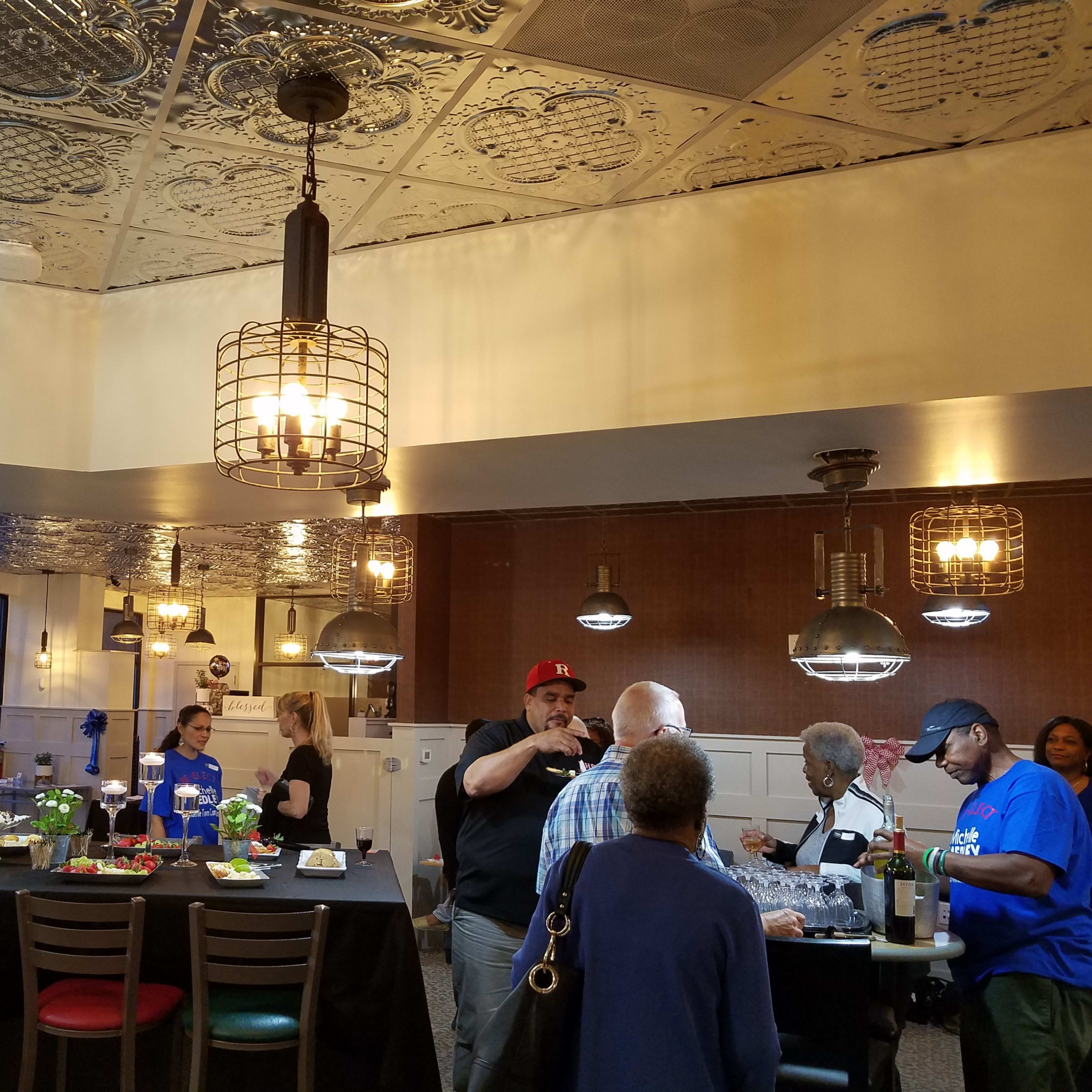 A group of people gathers in a well-lit restaurant with a central table adorned with food and drinks, while others engage in conversation.