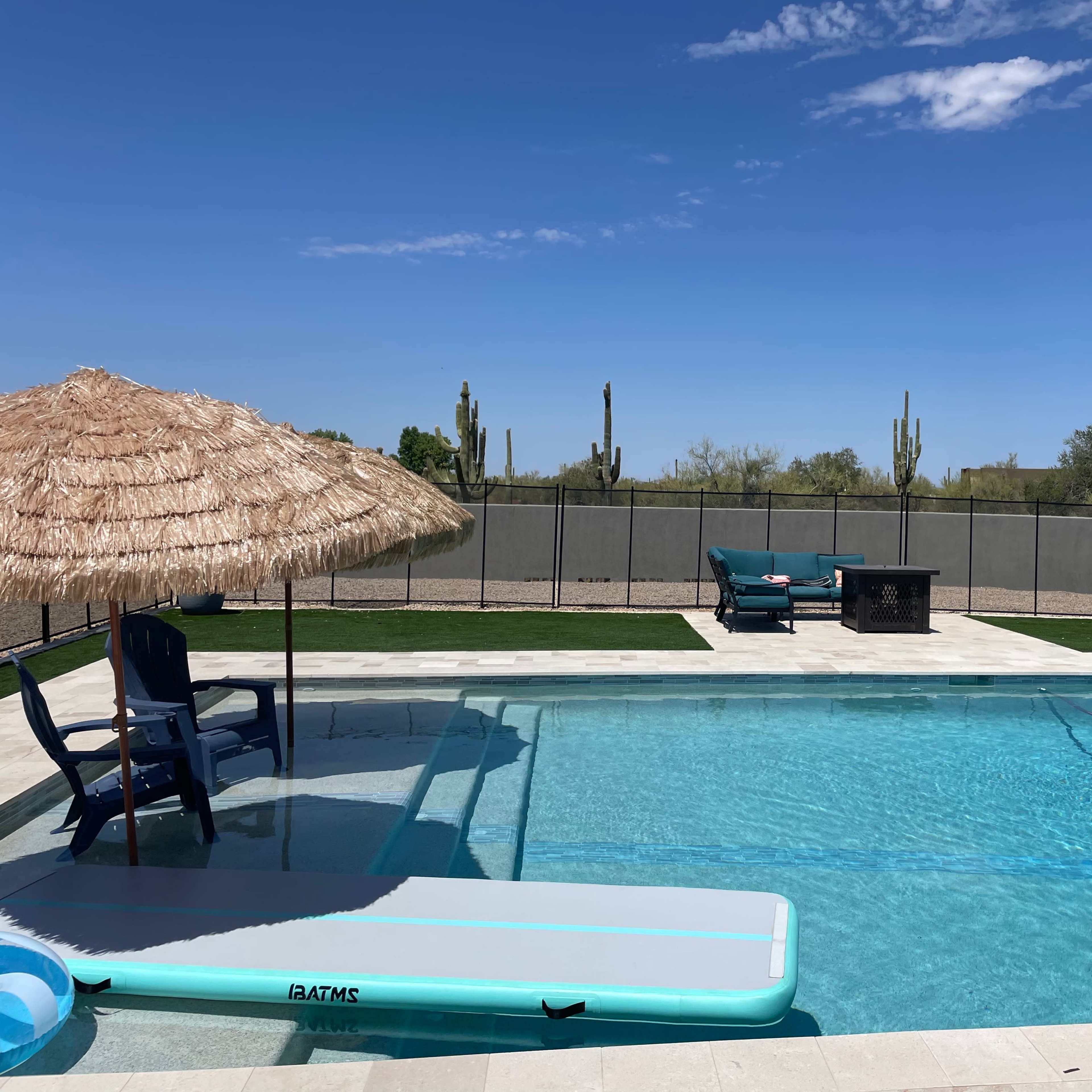 A swimming pool surrounded by a stone patio features a thatched umbrella, lounge chairs, and cacti in the background under a clear blue sky.