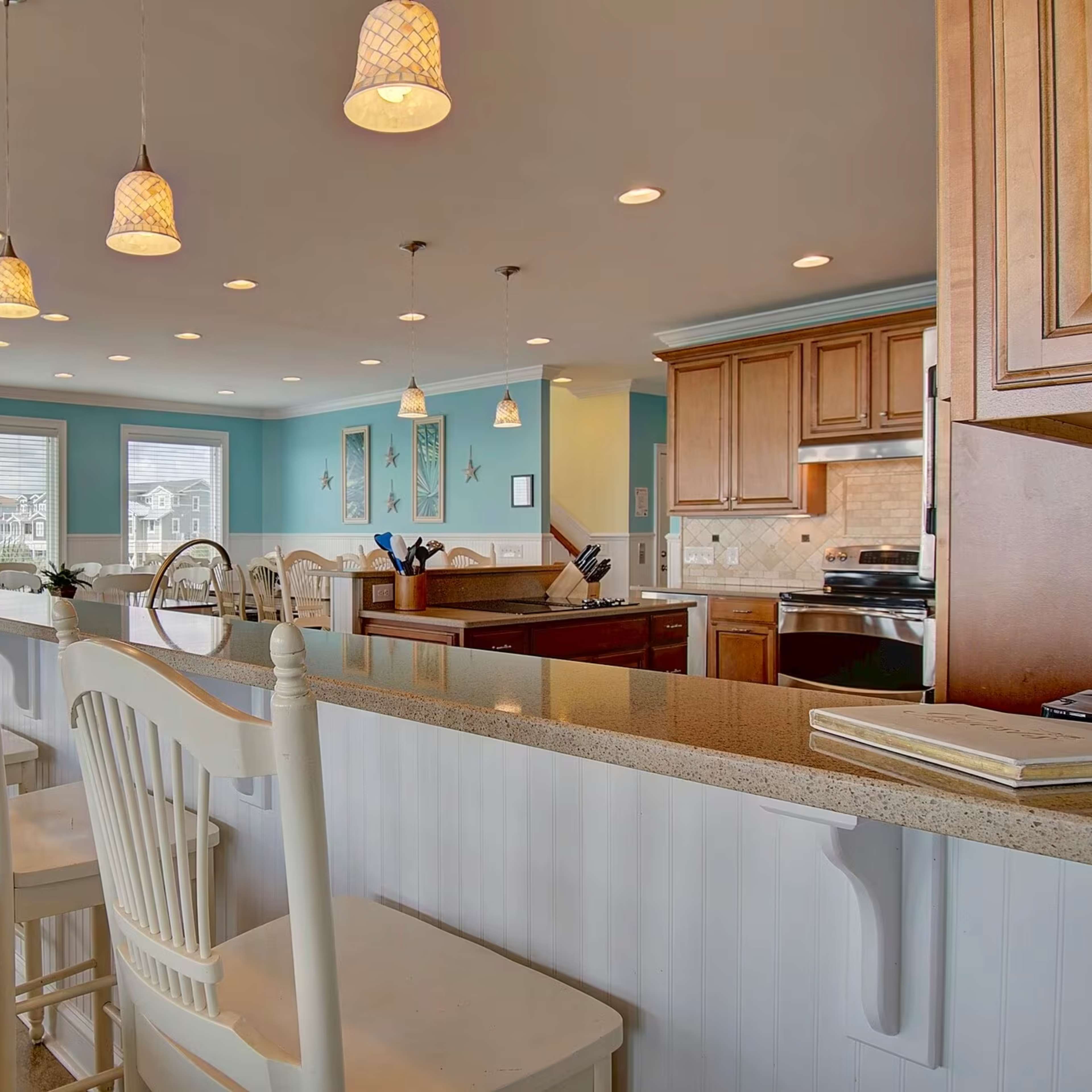 A spacious kitchen features a long bar with white stools, pendant lights, and a view of an adjacent dining area through large windows.