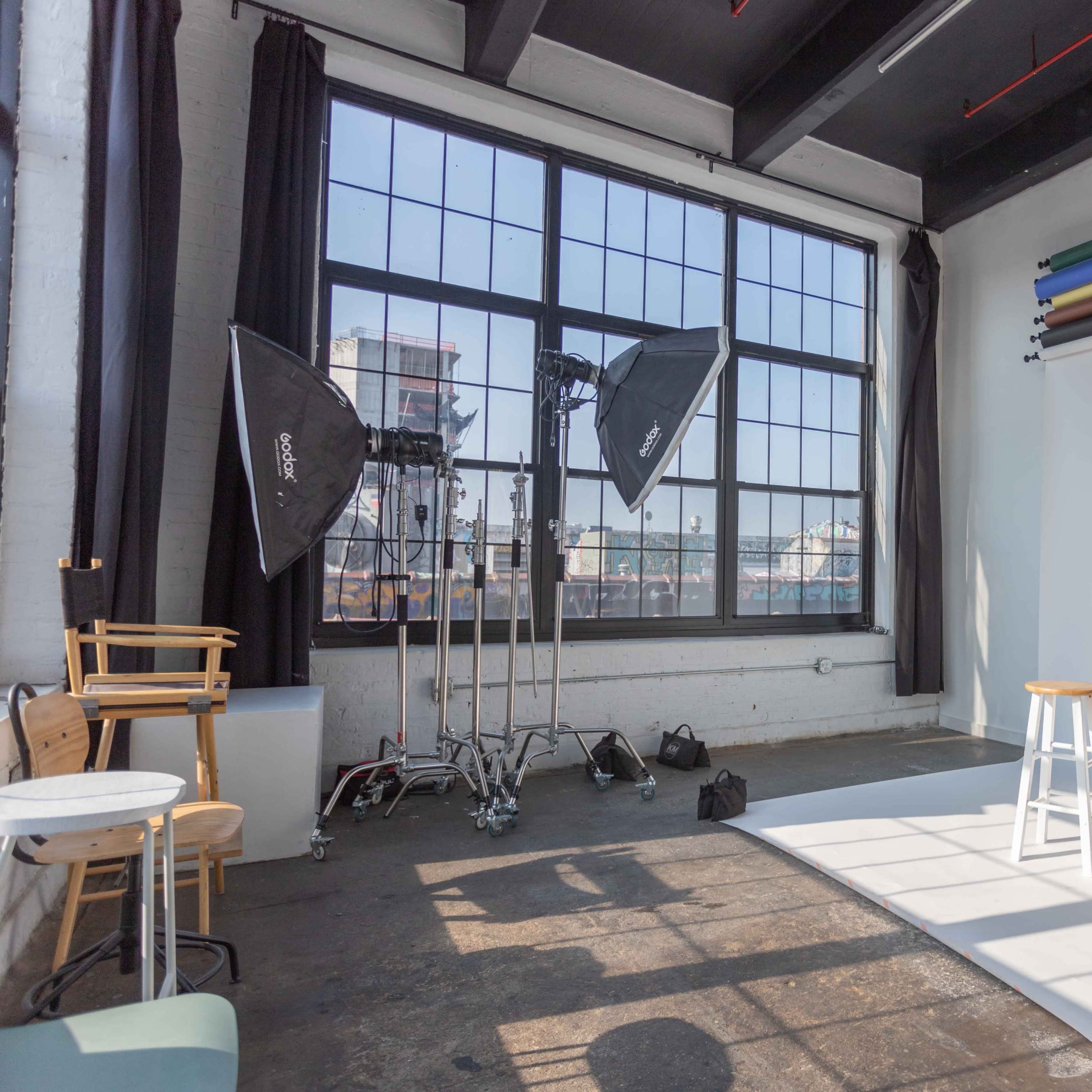 A photography studio featuring a white backdrop, a wooden stool, and several lighting equipment setups near large windows.
