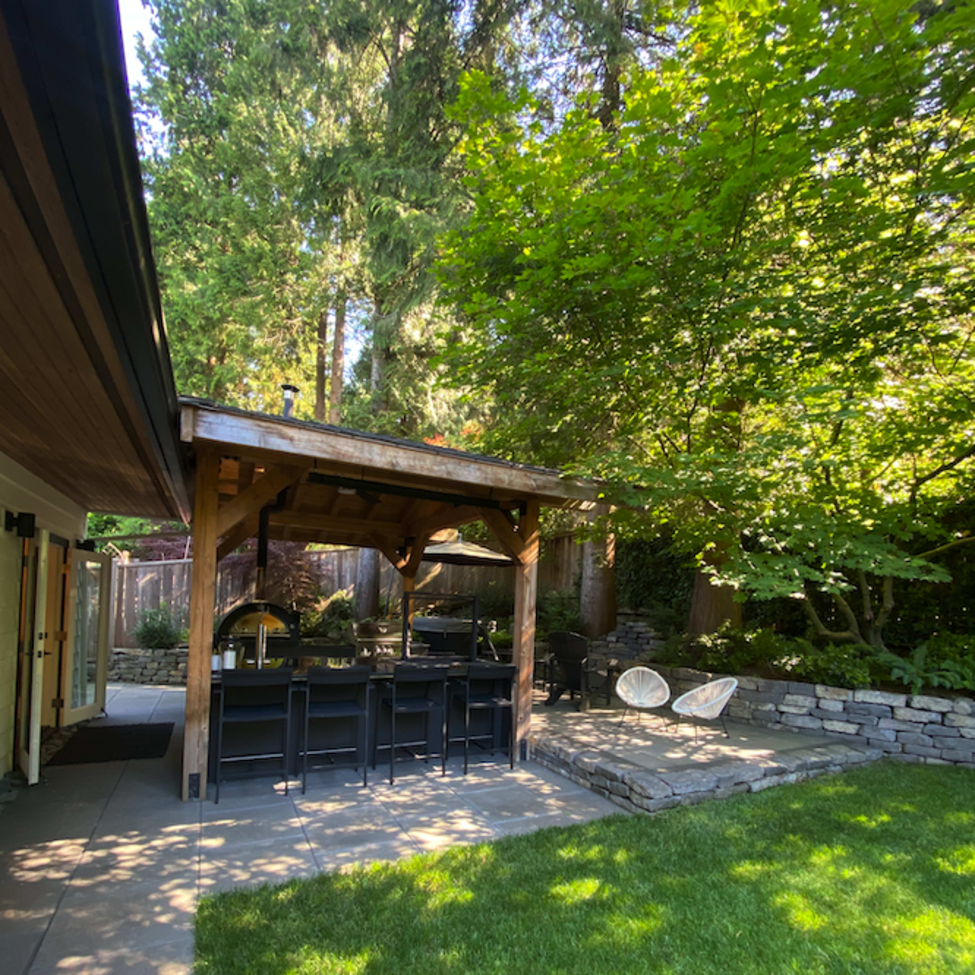 A wooden gazebo with seating is positioned next to a grassy area surrounded by trees and stone landscaping.