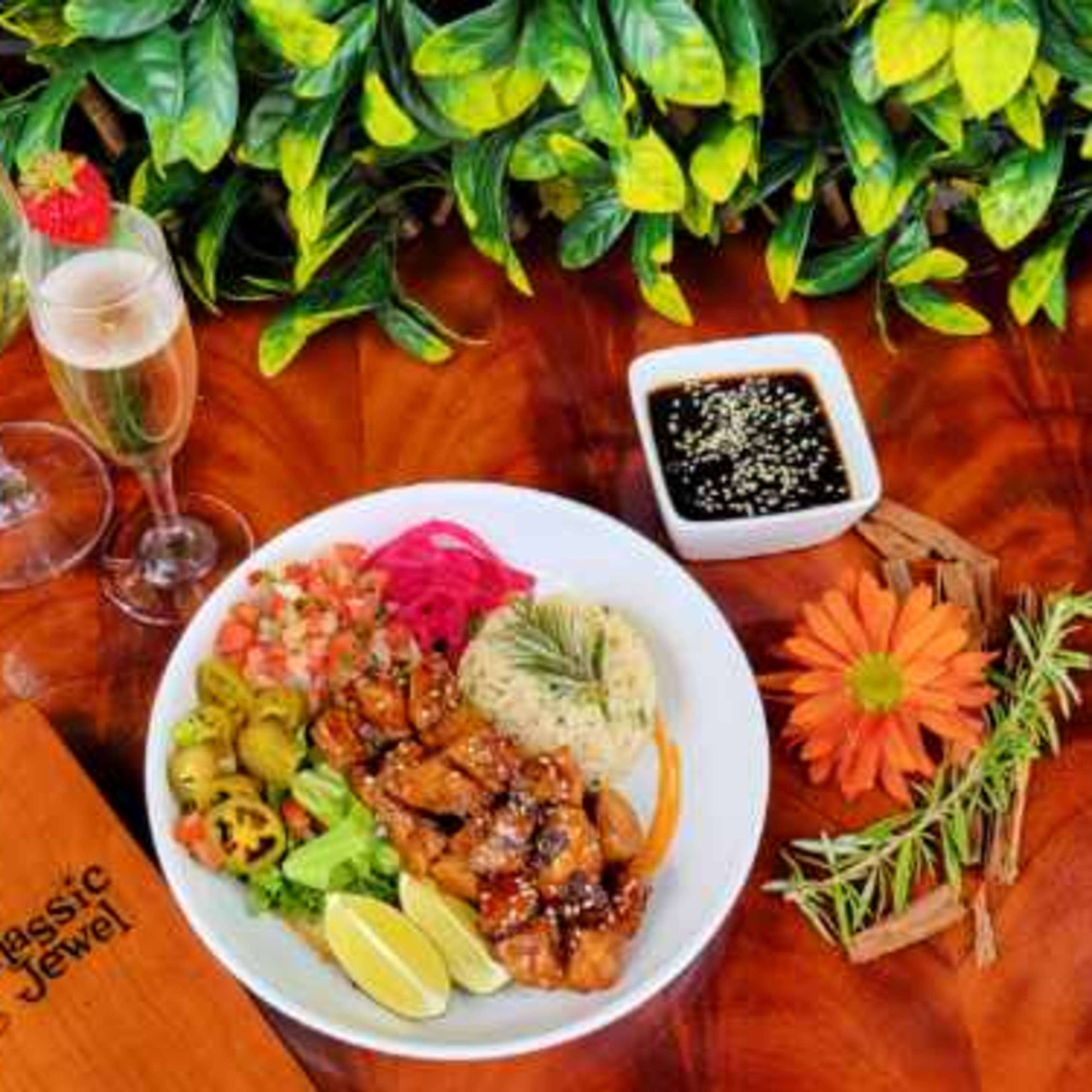 A plated meal with grilled chicken, rice, vegetables, and sauces, accompanied by two drinks and garnished with a flower on a wooden table.
