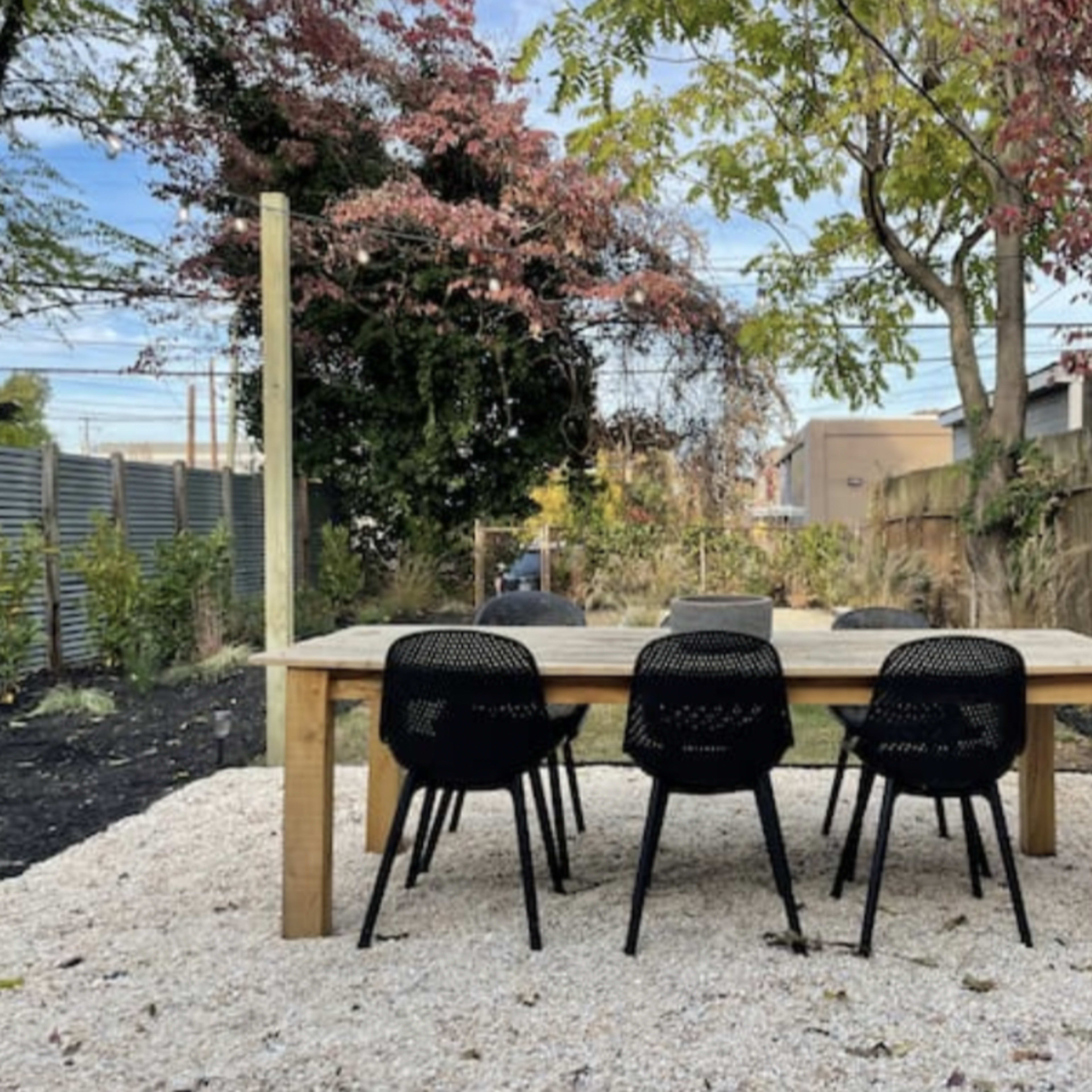 A wooden dining table surrounded by black chairs sits on a gravel surface in a garden with trees and shrubs.