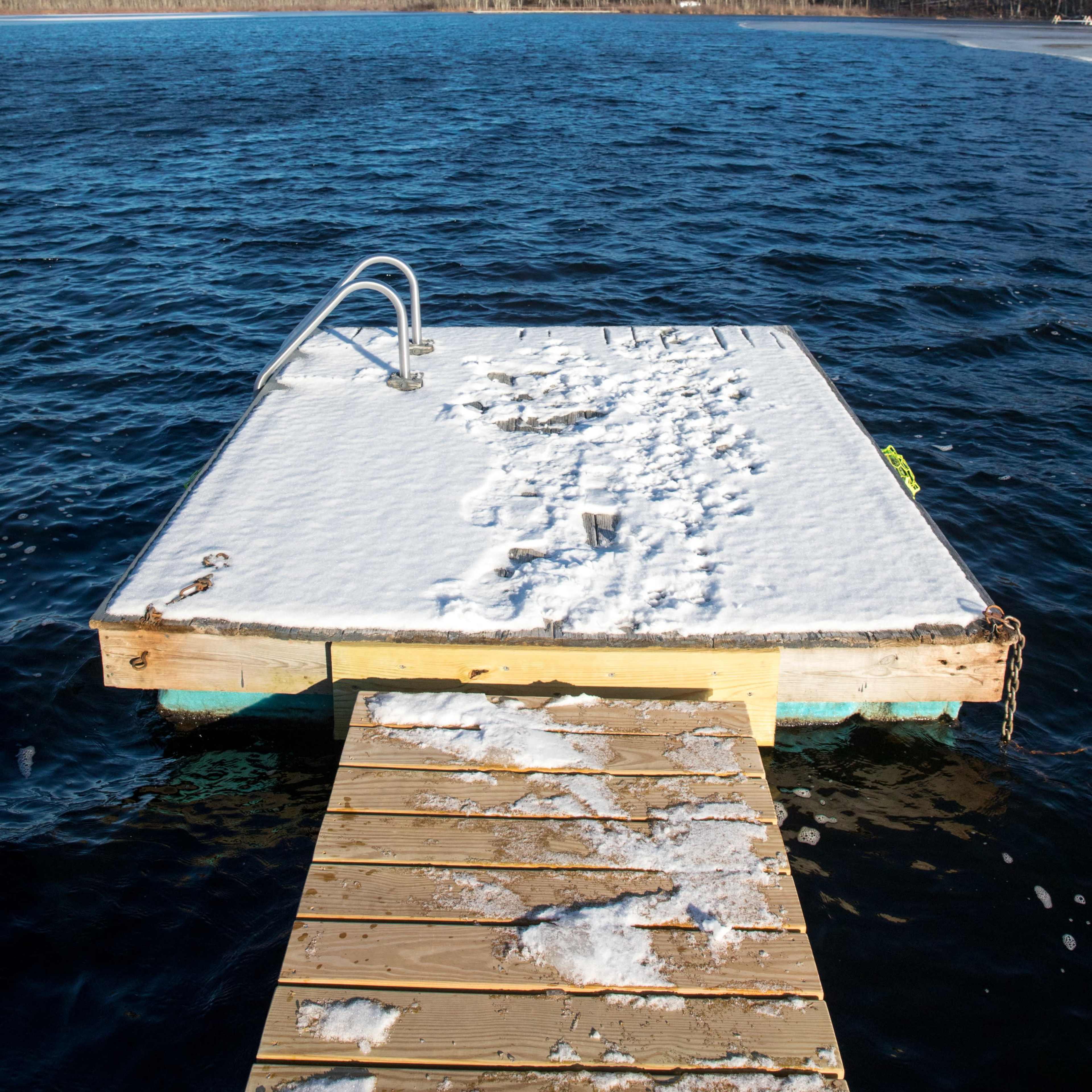 A snow-covered dock extends into a lake surrounded by a treeline under a clear blue sky.