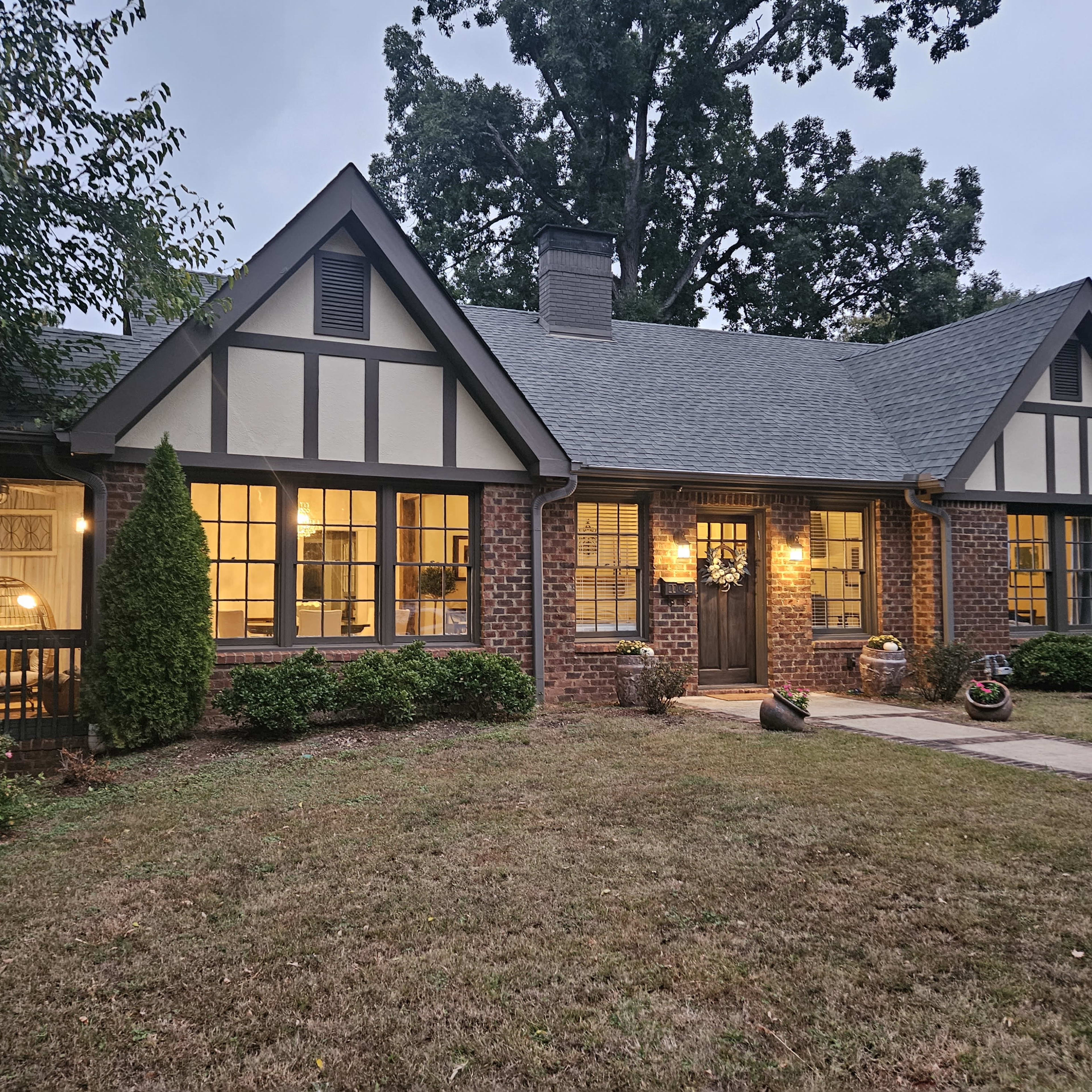 A Tudor-style brick house with a steeply pitched roof and large windows, surrounded by greenery and a neatly maintained lawn.