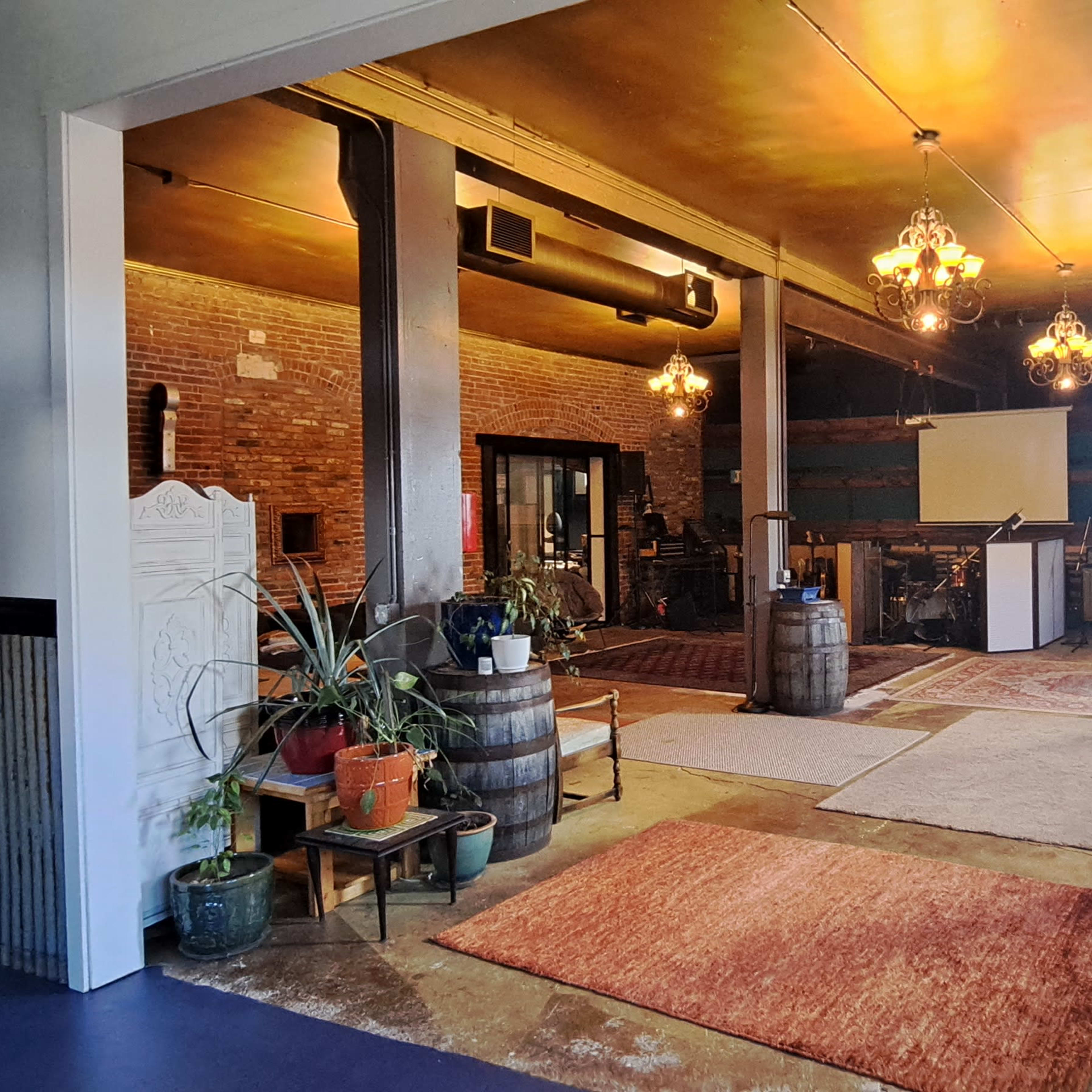 A spacious, well-lit room with exposed brick walls, wooden beams, and various plants and decor items, featuring rugs and barrels as furnishings.