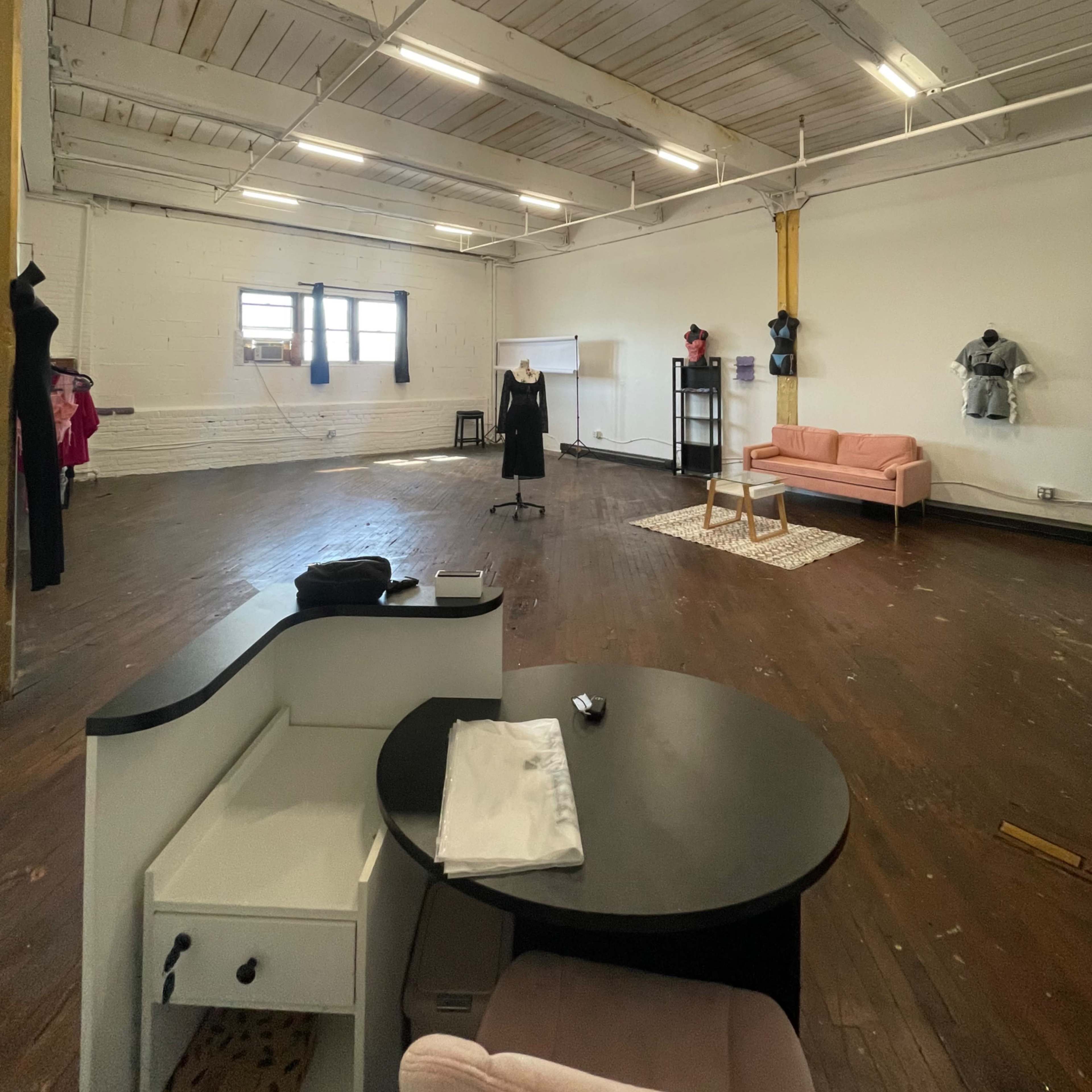 A spacious, empty room with wooden flooring, featuring a small black table, a pink sofa, and mannequins dressed in various outfits.