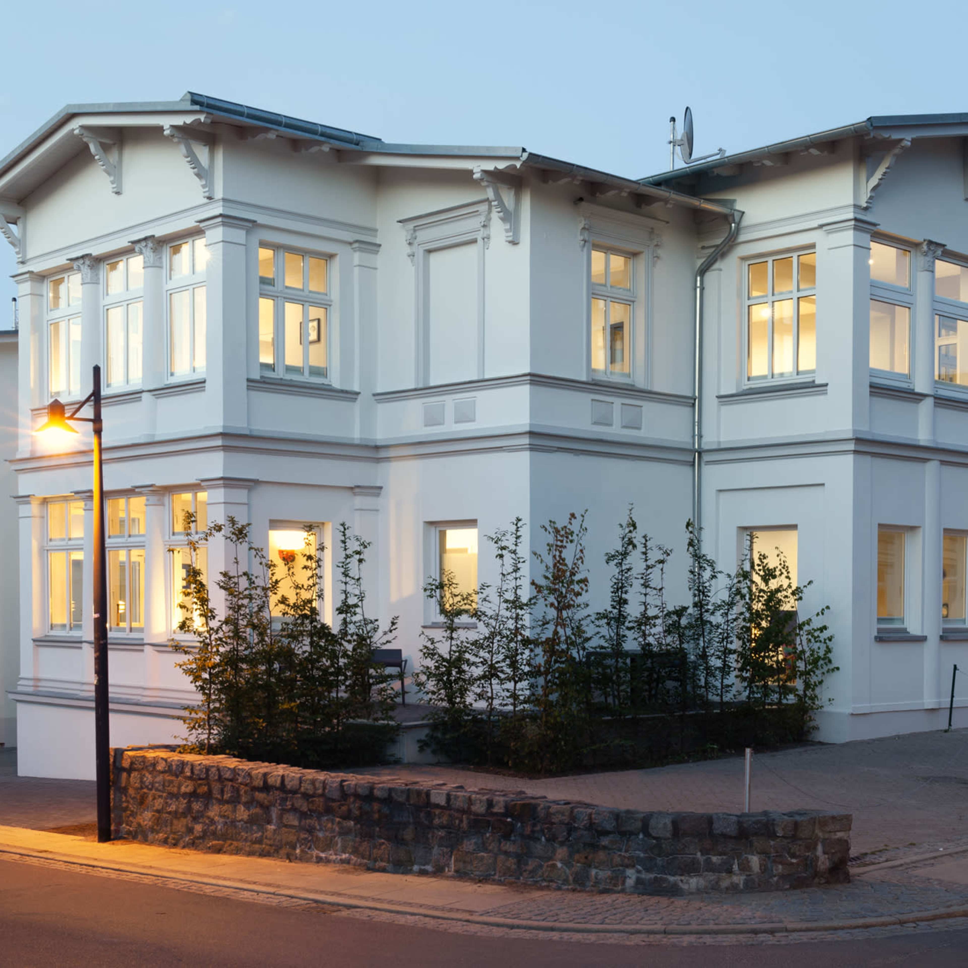 A two-story white house with large windows and a stone wall in front, situated at the corner of a street during twilight.