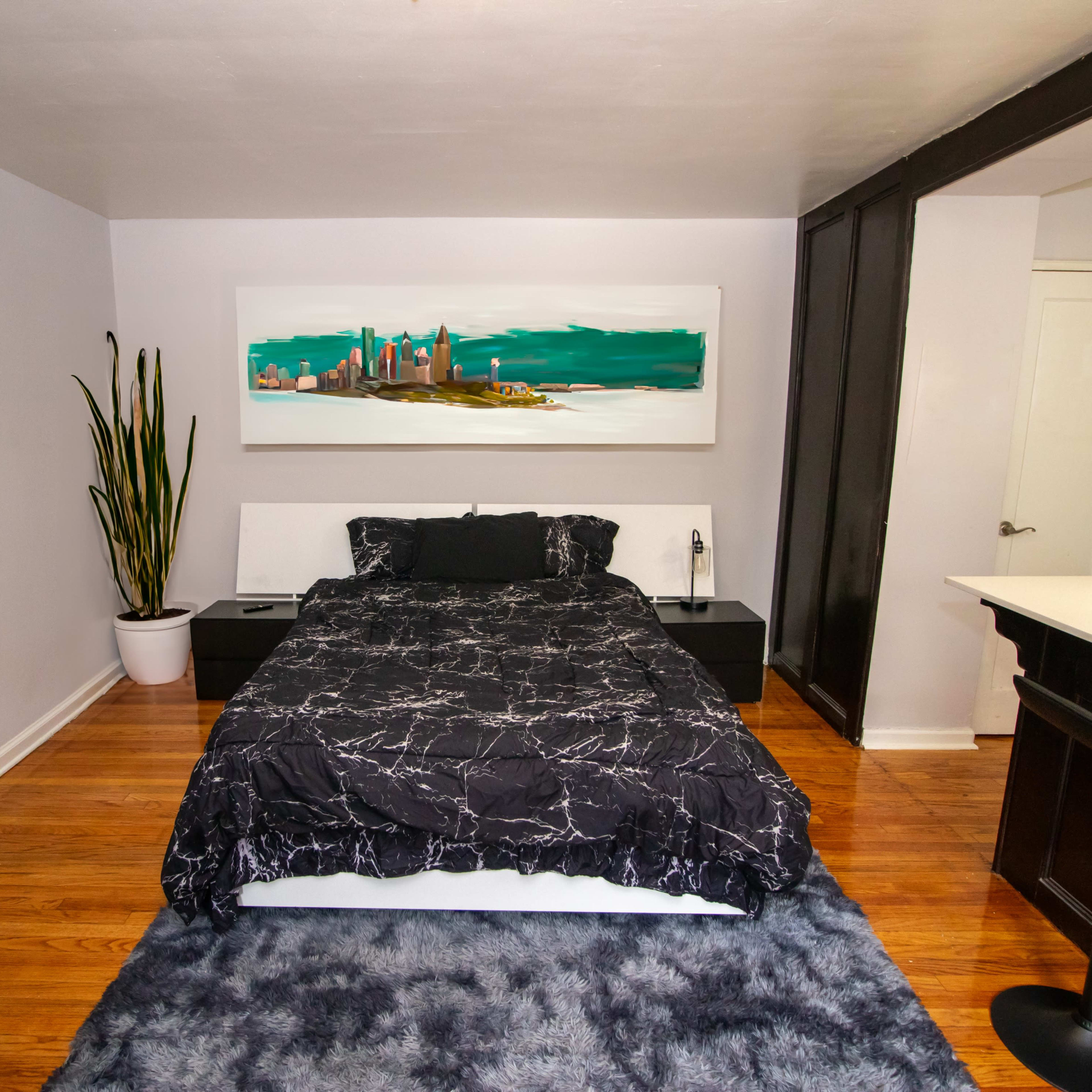 A bedroom features a black and white marble-patterned bedspread, a large painting of a cityscape above the bed, and a potted plant in the corner.