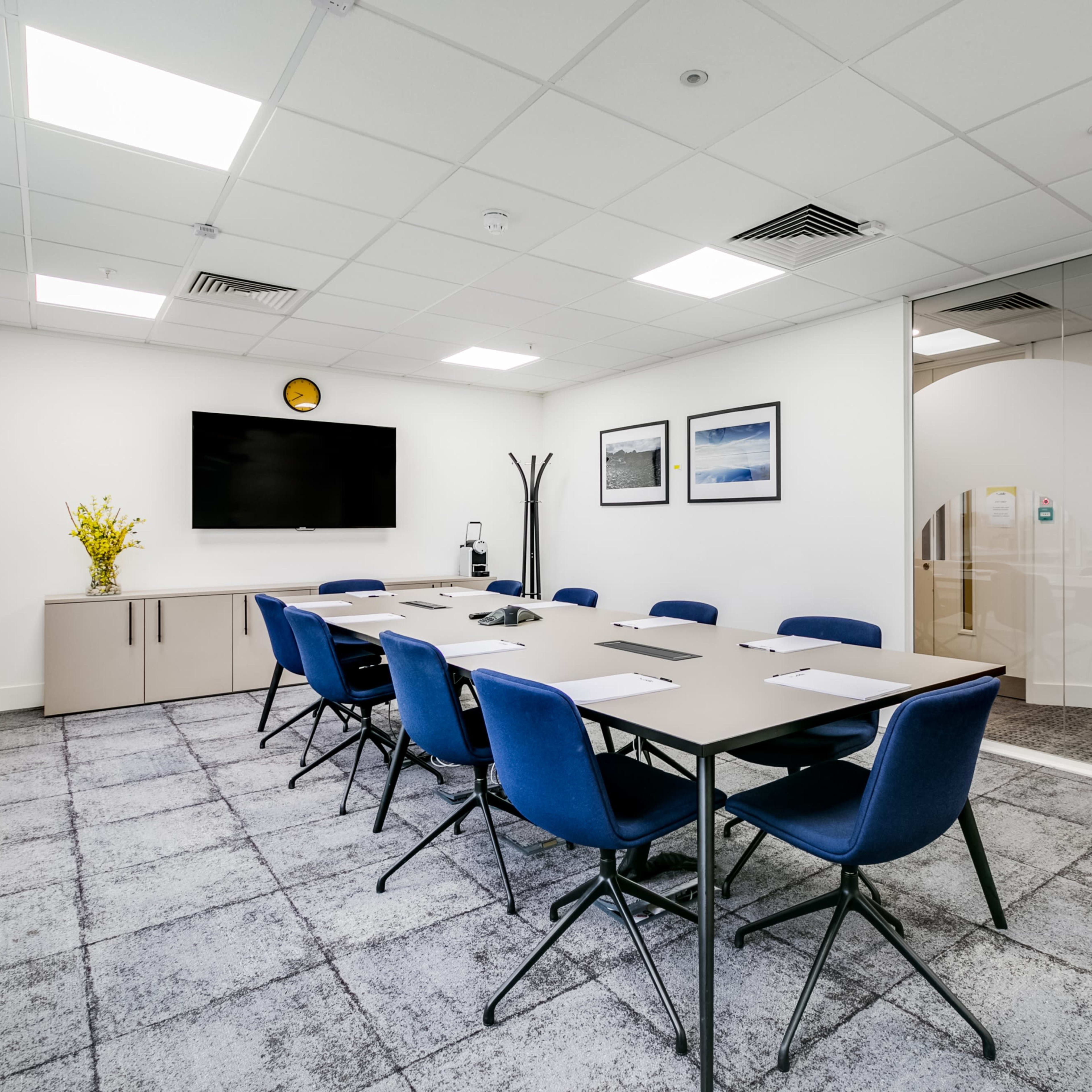 A well-lit conference room features a long table surrounded by blue chairs, a large screen on the wall, and glass partitions.