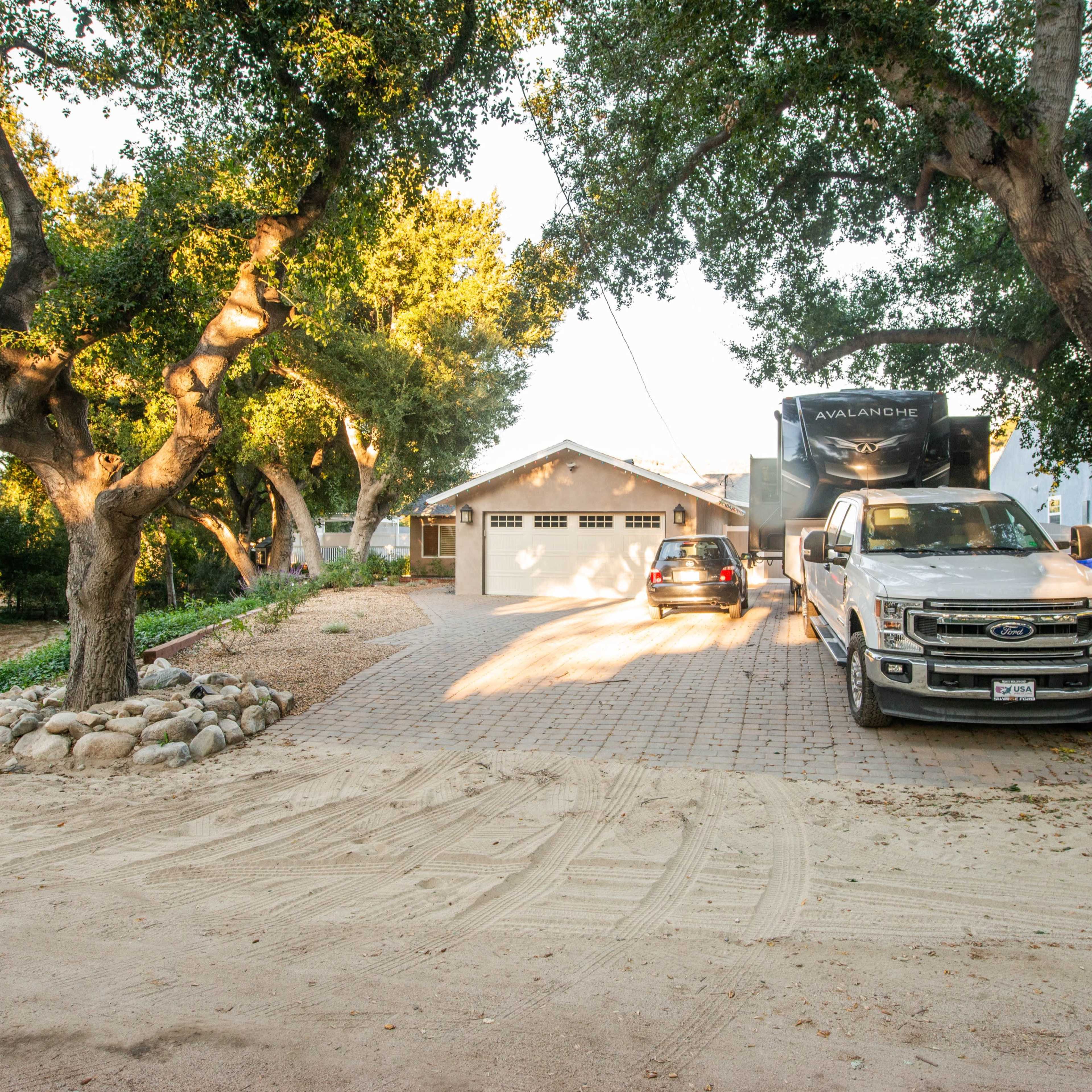 A gravel driveway leads to a house surrounded by trees, with two parked vehicles, including a truck and an RV.