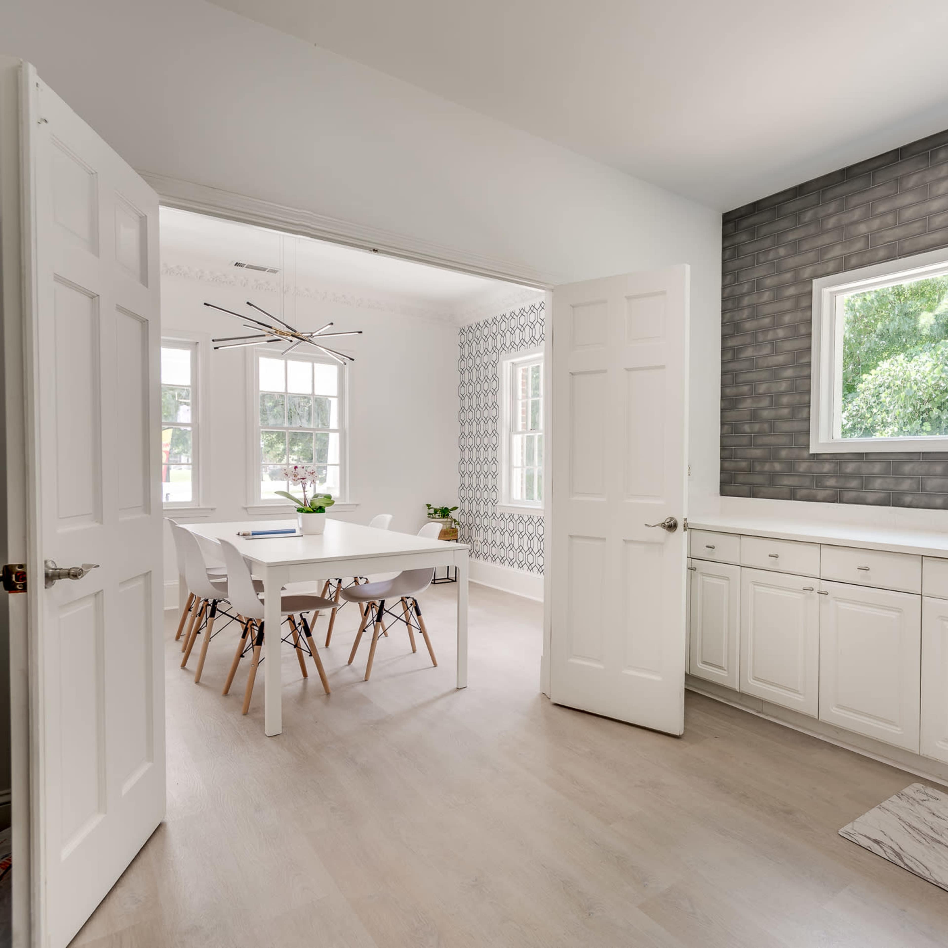 A modern kitchen features white cabinets and a window with a view of greenery, adjacent to a dining area with a large table and stylish chairs.