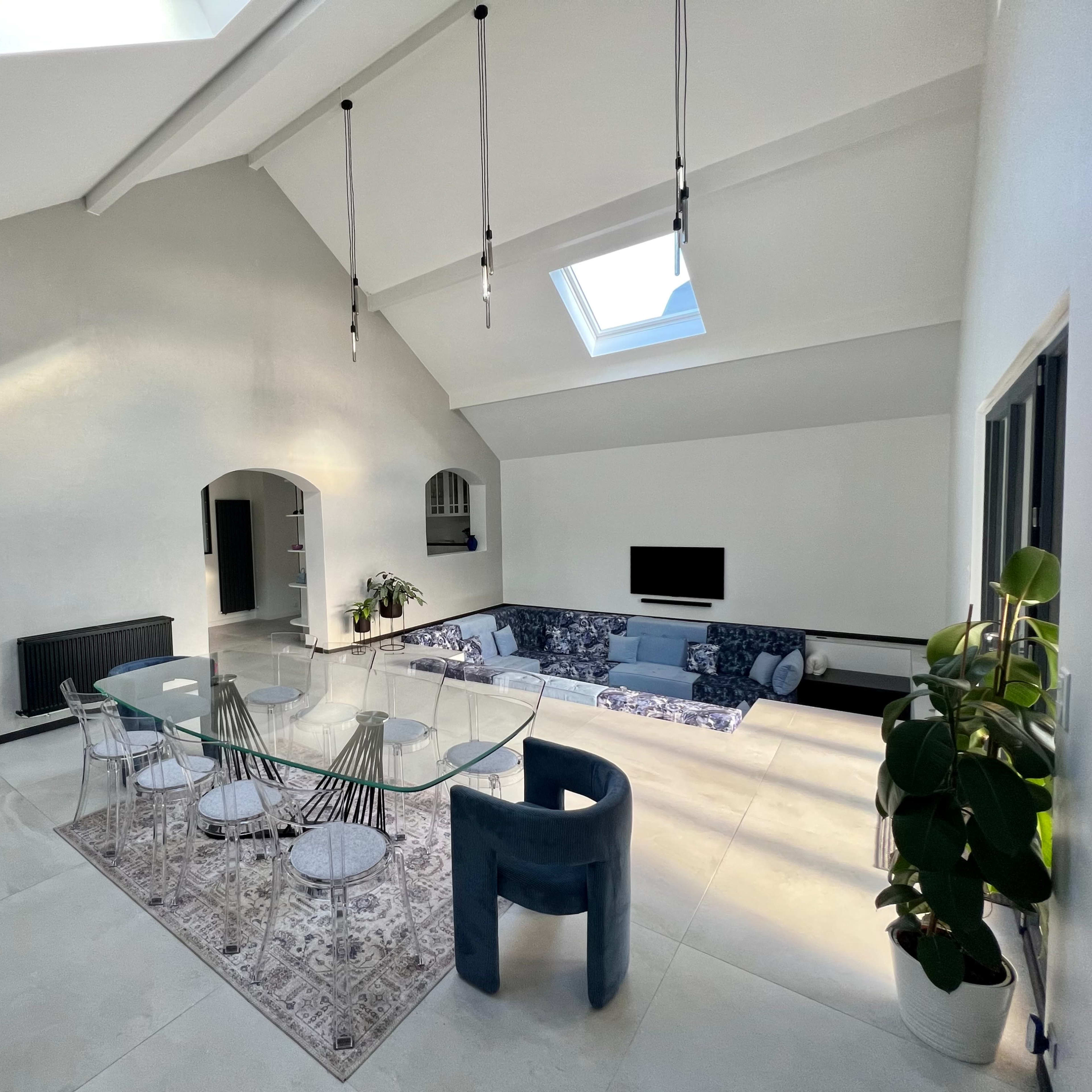 A spacious dining area features a glass table surrounded by transparent chairs, with a skylight illuminating the room and a modern sectional sofa in the background.