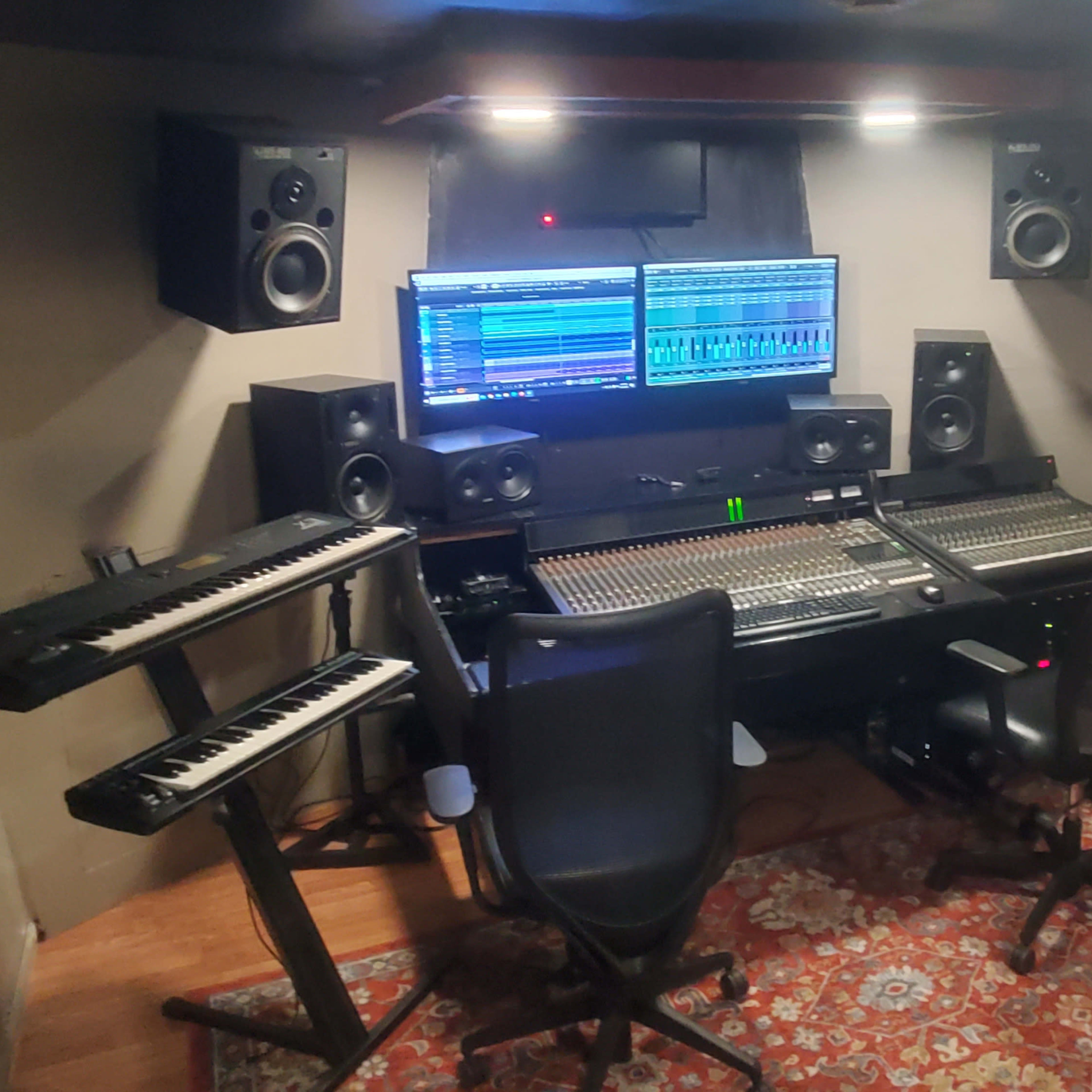 A music recording studio featuring mixing equipment, two computer monitors, audio speakers, and synthesizers arranged around a central mixing console.