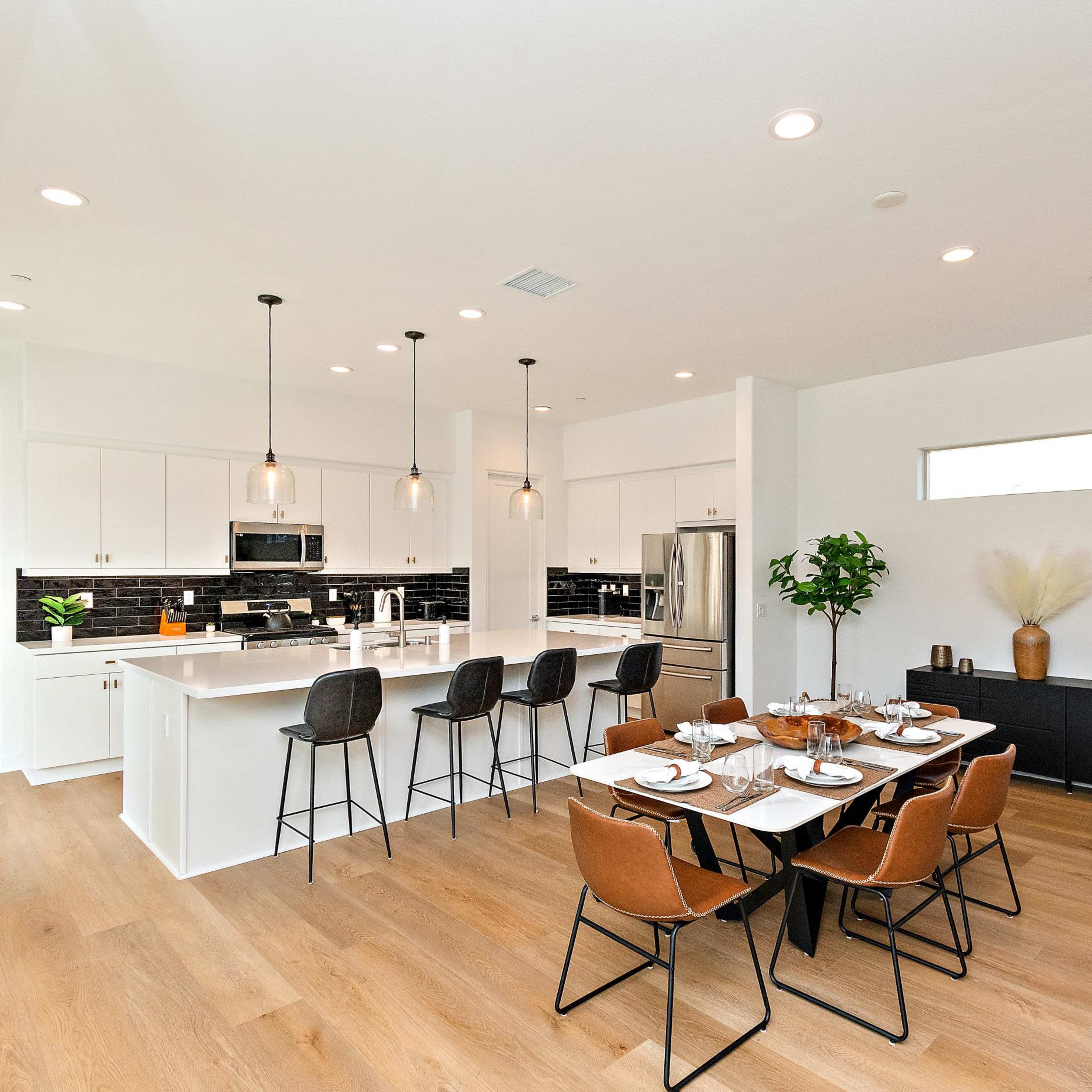 A modern kitchen and dining area features white cabinetry, black tile backsplash, and a large dining table set with dishes, surrounded by stylish chairs.