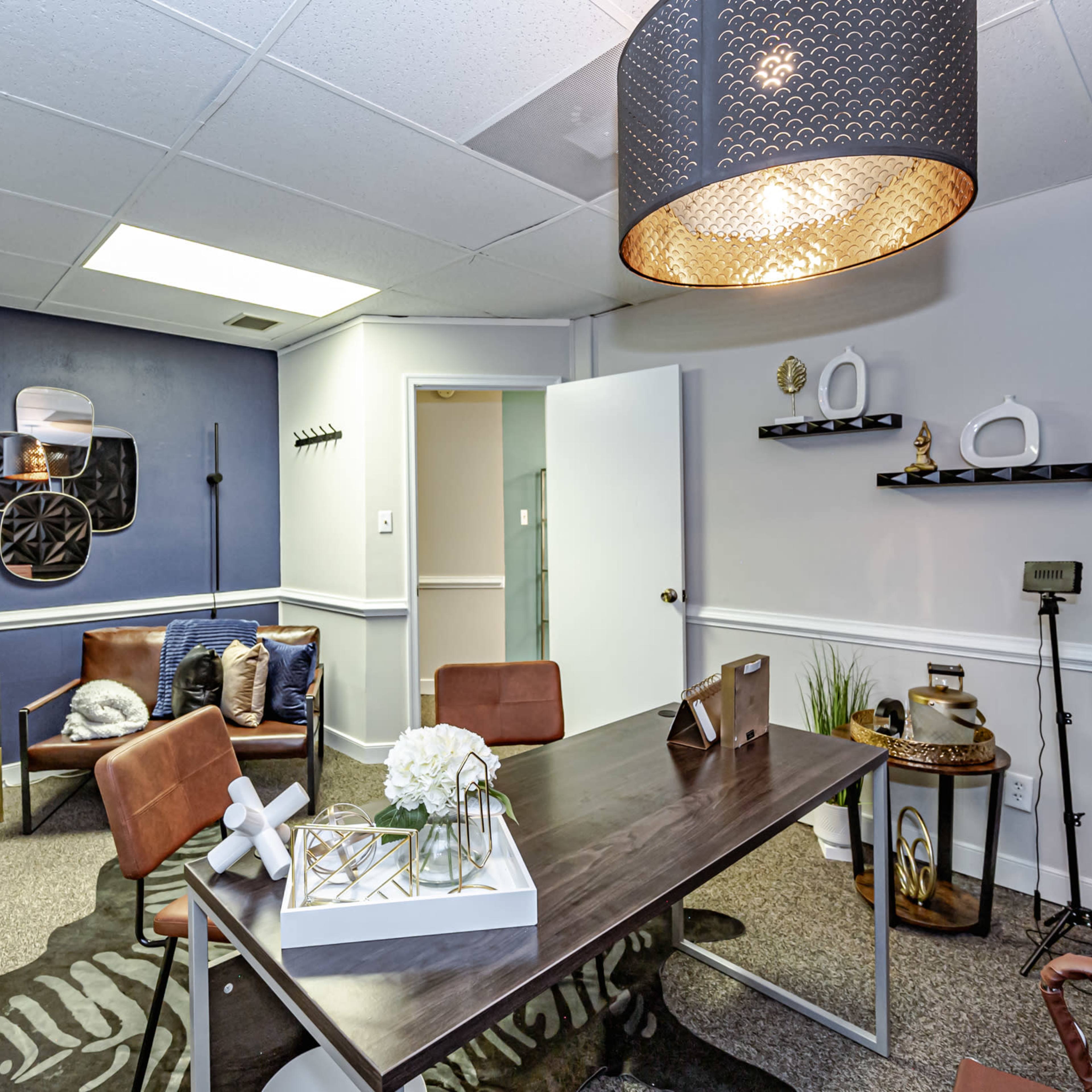 A modern office space features a table, chairs, decorative items, and a large overhead light, with dark blue walls and carpeting.