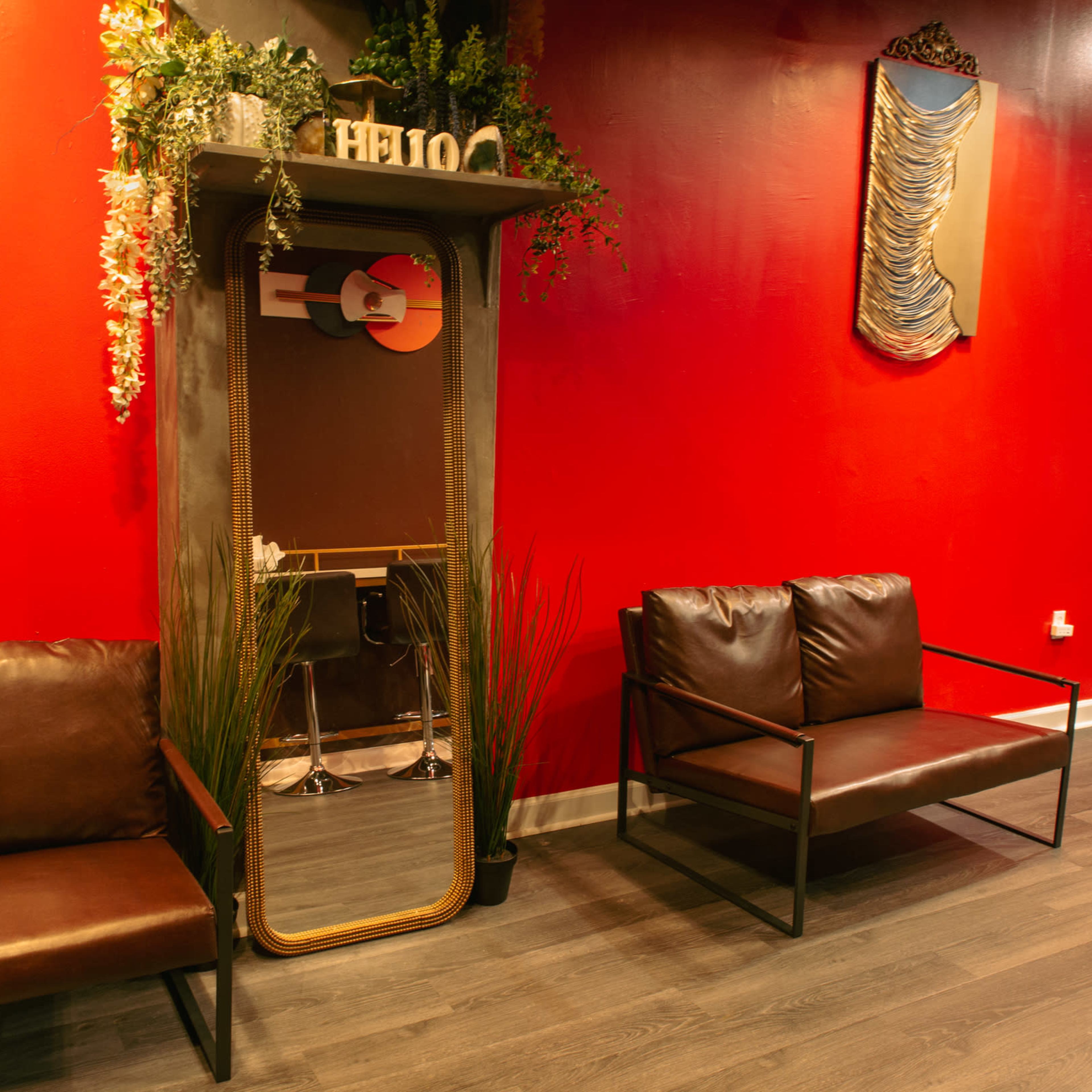 A waiting area with two brown couches, a mirror framed by greenery, and a striking red wall in the background.