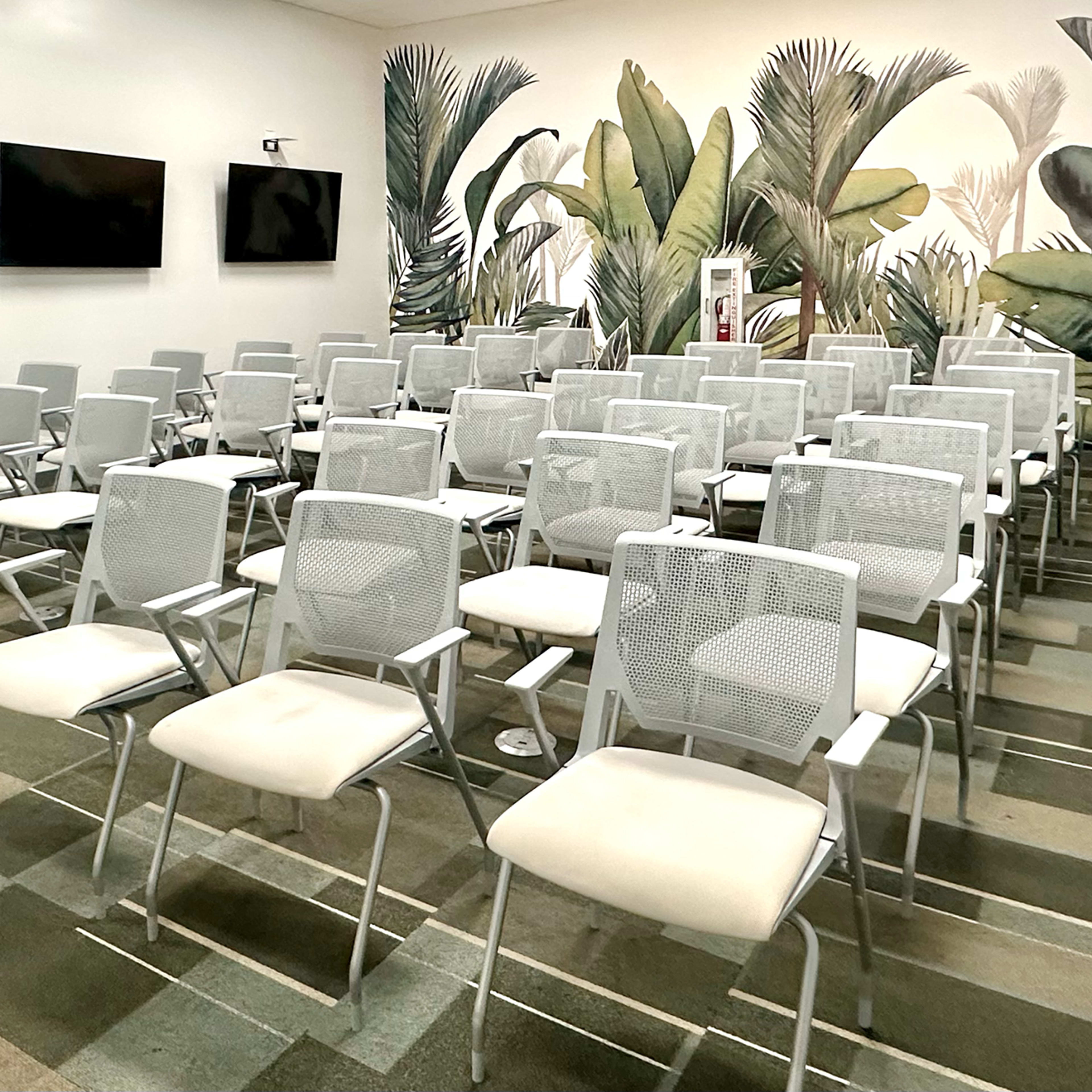A conference room is arranged with multiple rows of white chairs facing a wall adorned with leafy murals and equipped with two televisions.