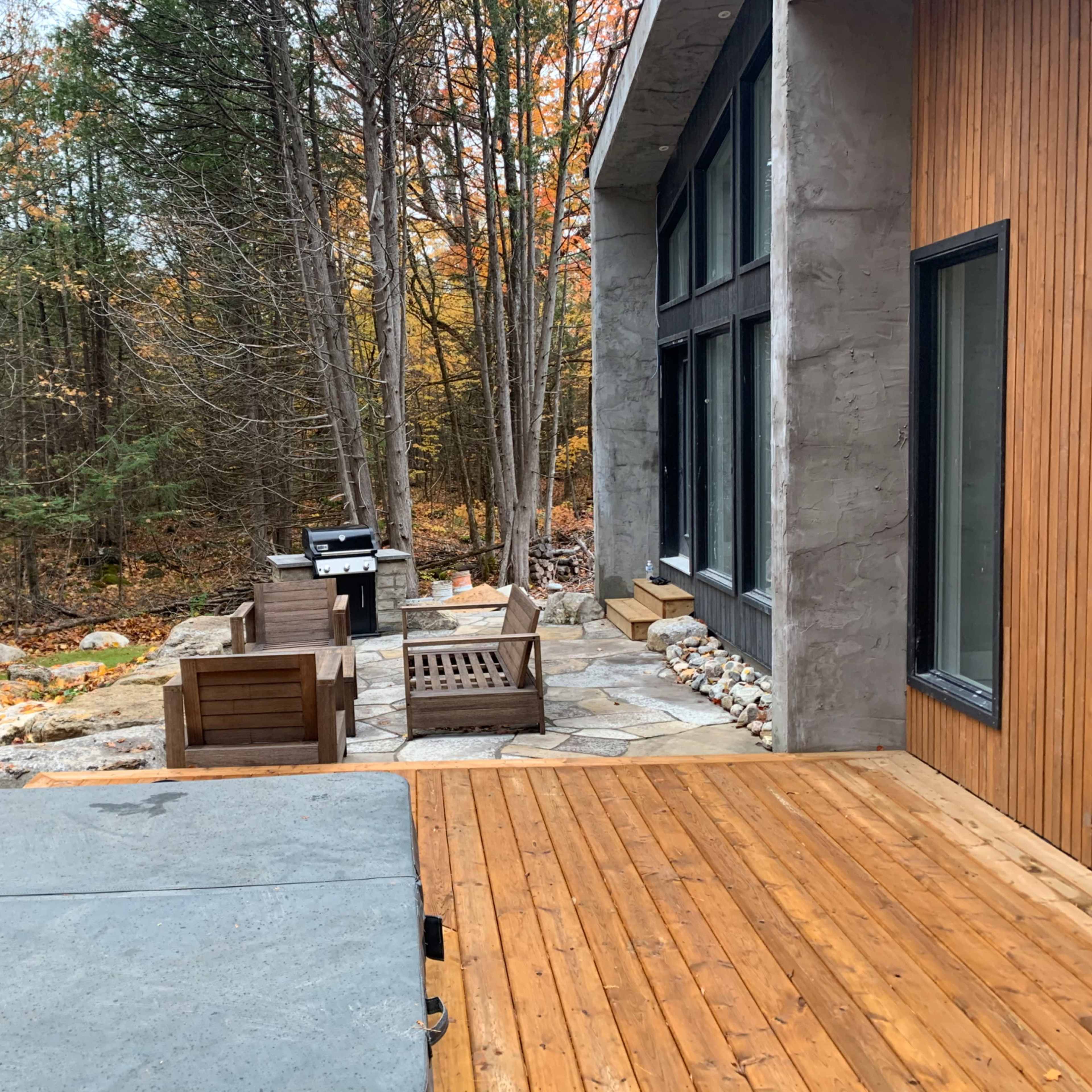 A wooden deck extends from a modern house, leading to a stone patio with outdoor seating and a grill, surrounded by trees displaying autumn foliage.