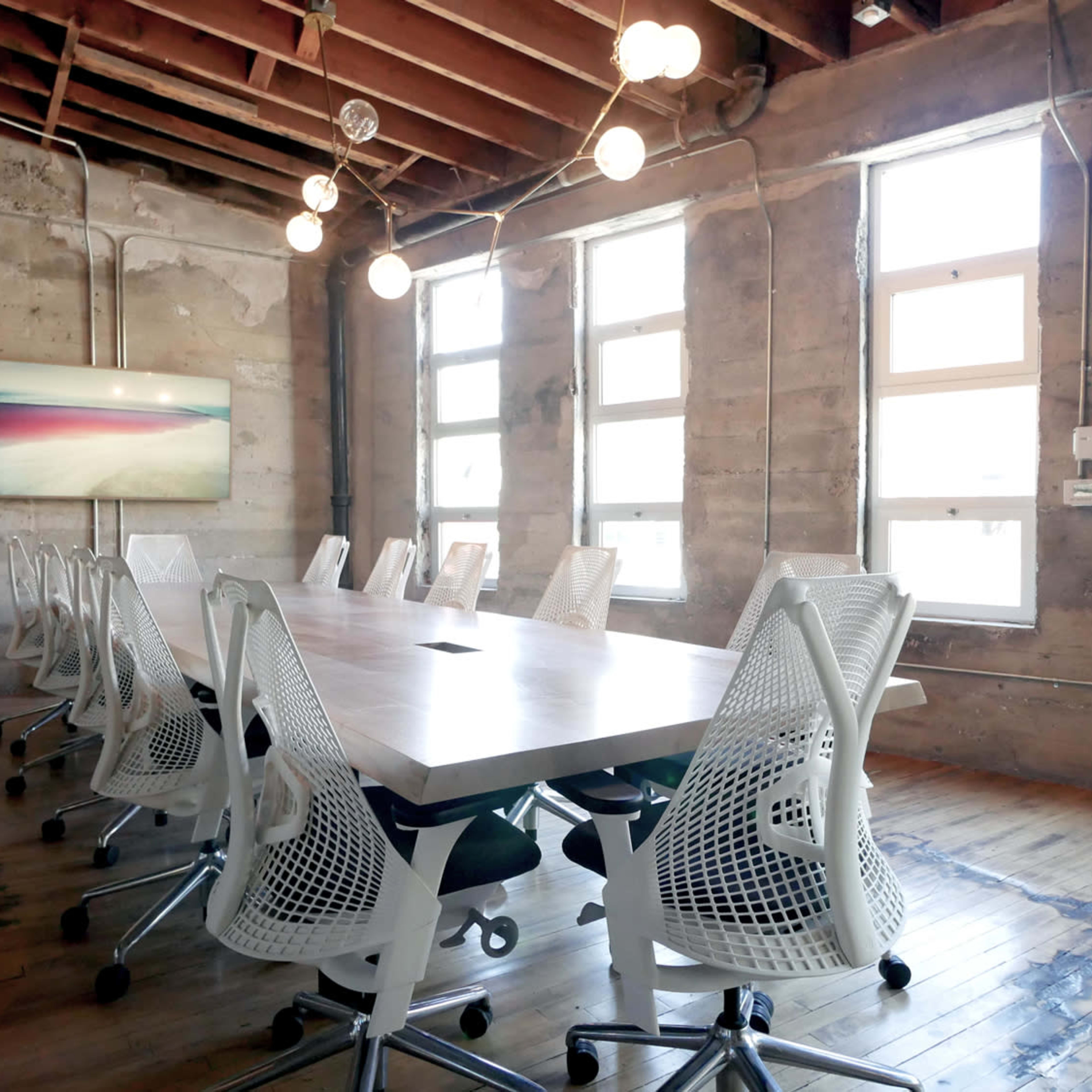 A long conference table with white mesh chairs is set up in a brightly lit room featuring exposed wooden beams and concrete walls.