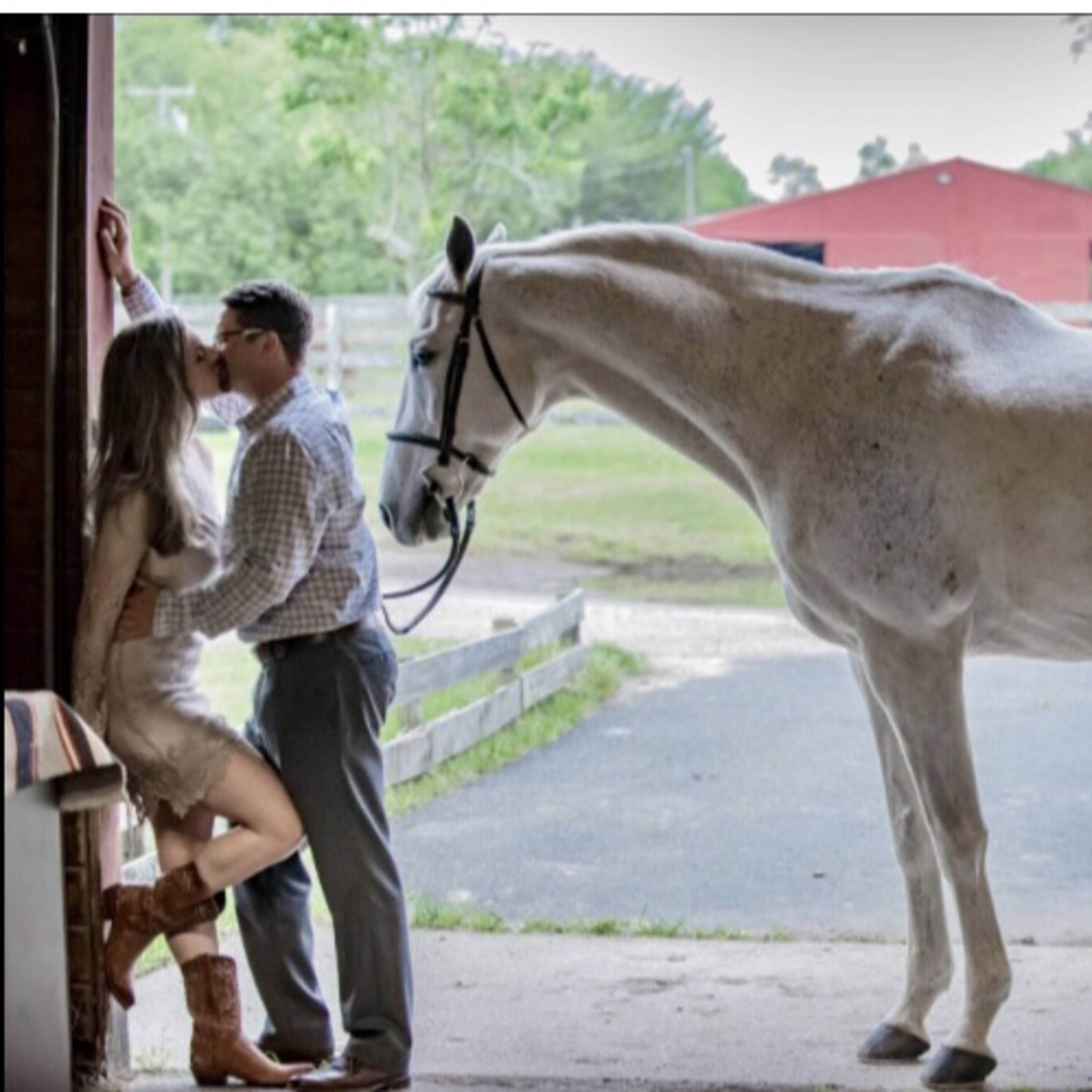 A couple kisses near the entrance of a stable while a white horse stands nearby.