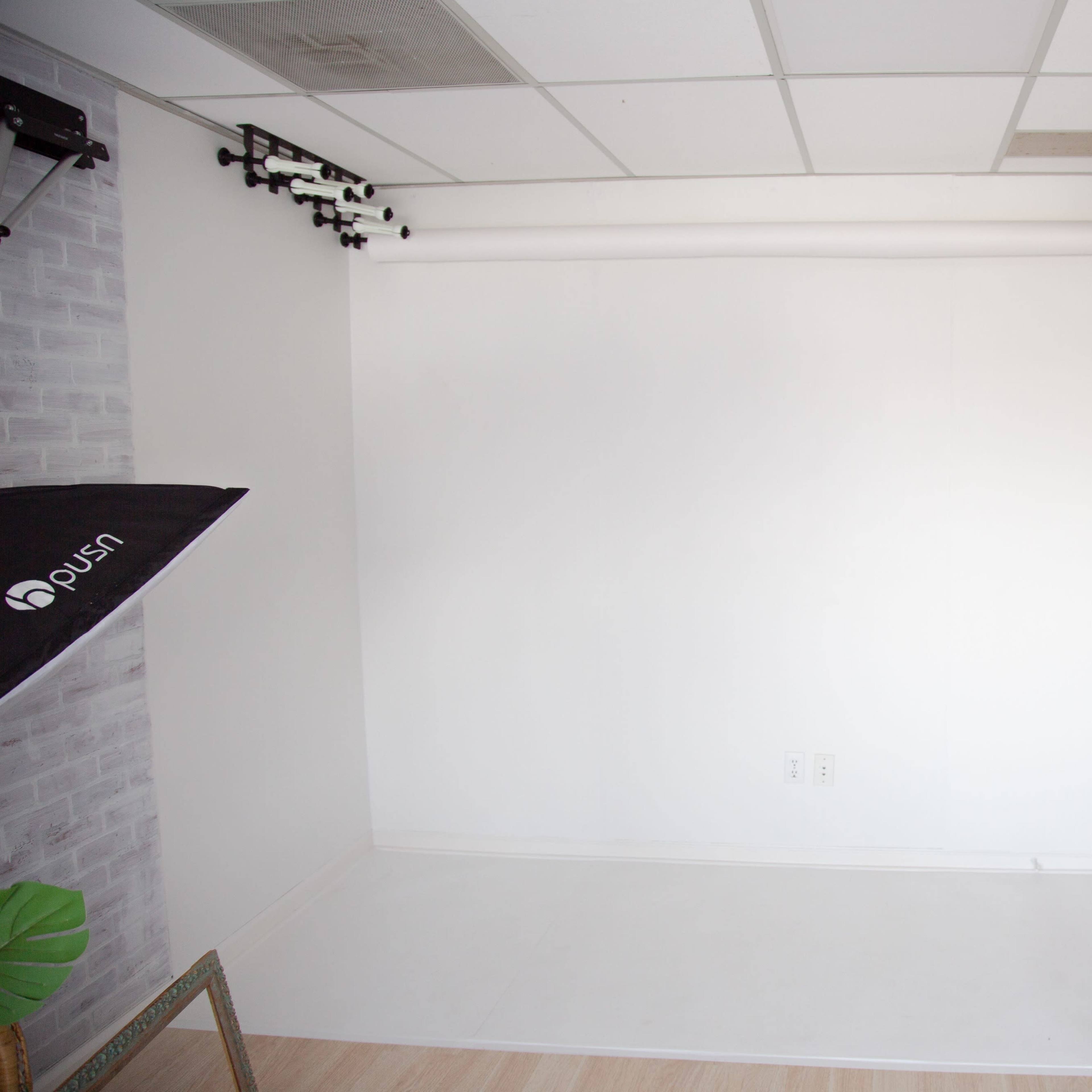 A minimalistic studio space with a white backdrop, a door on the right, and lighting equipment mounted on the ceiling.