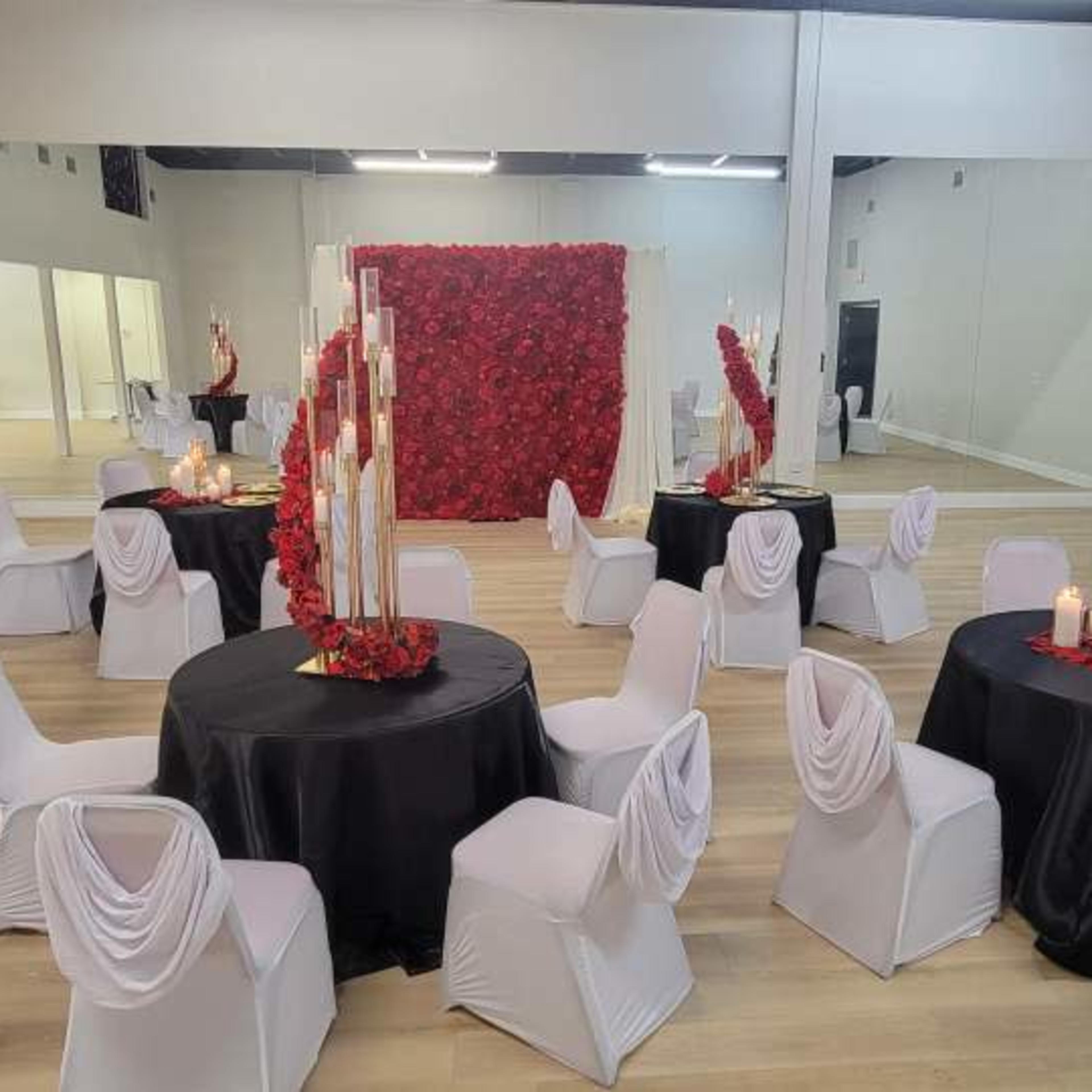 A spacious event venue arranged with round tables covered in black cloth, white chairs with draped backs, and a backdrop of red roses.