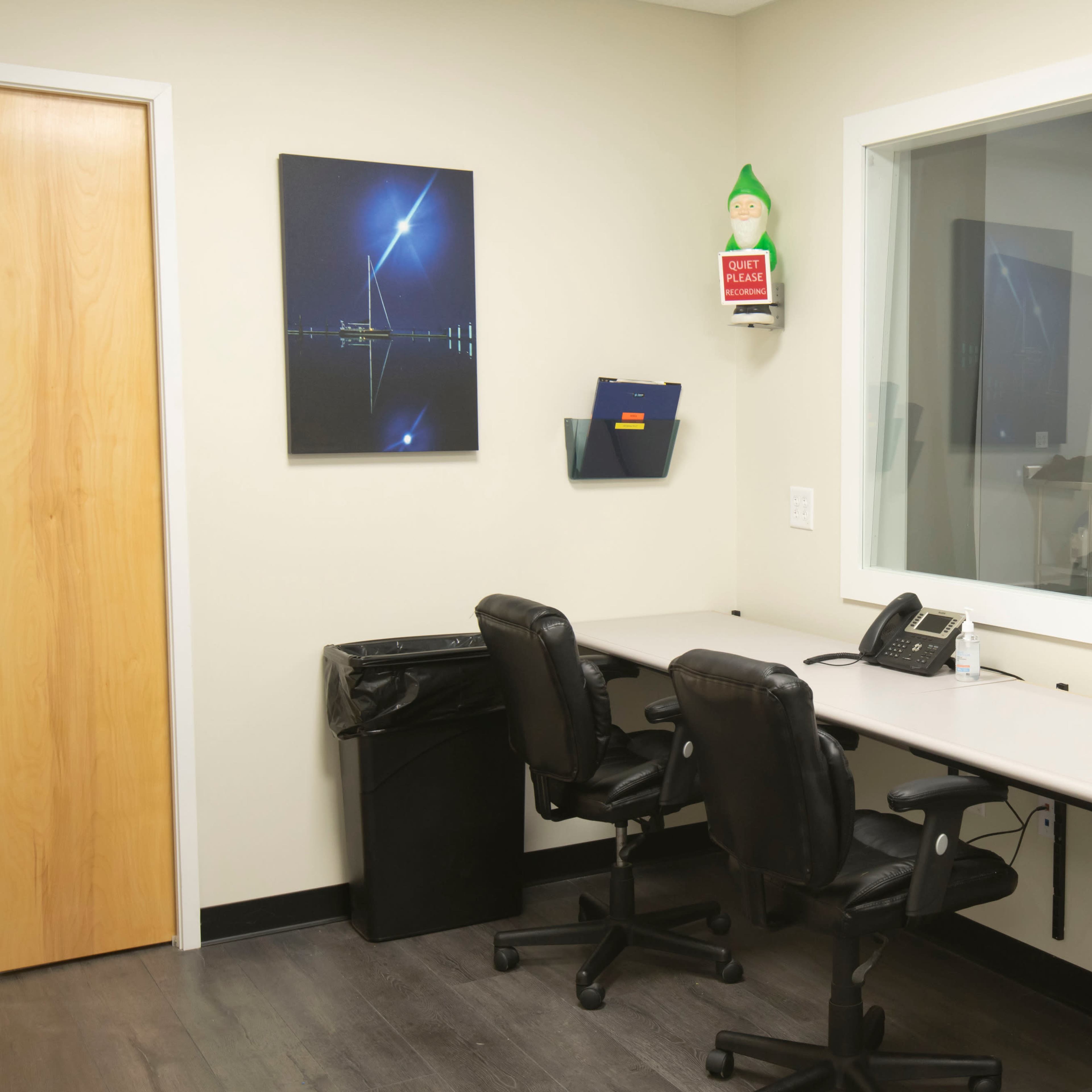 A small office space with two black chairs positioned at a gray desk, a telephone, a trash can, and a window with a framed picture on the wall.