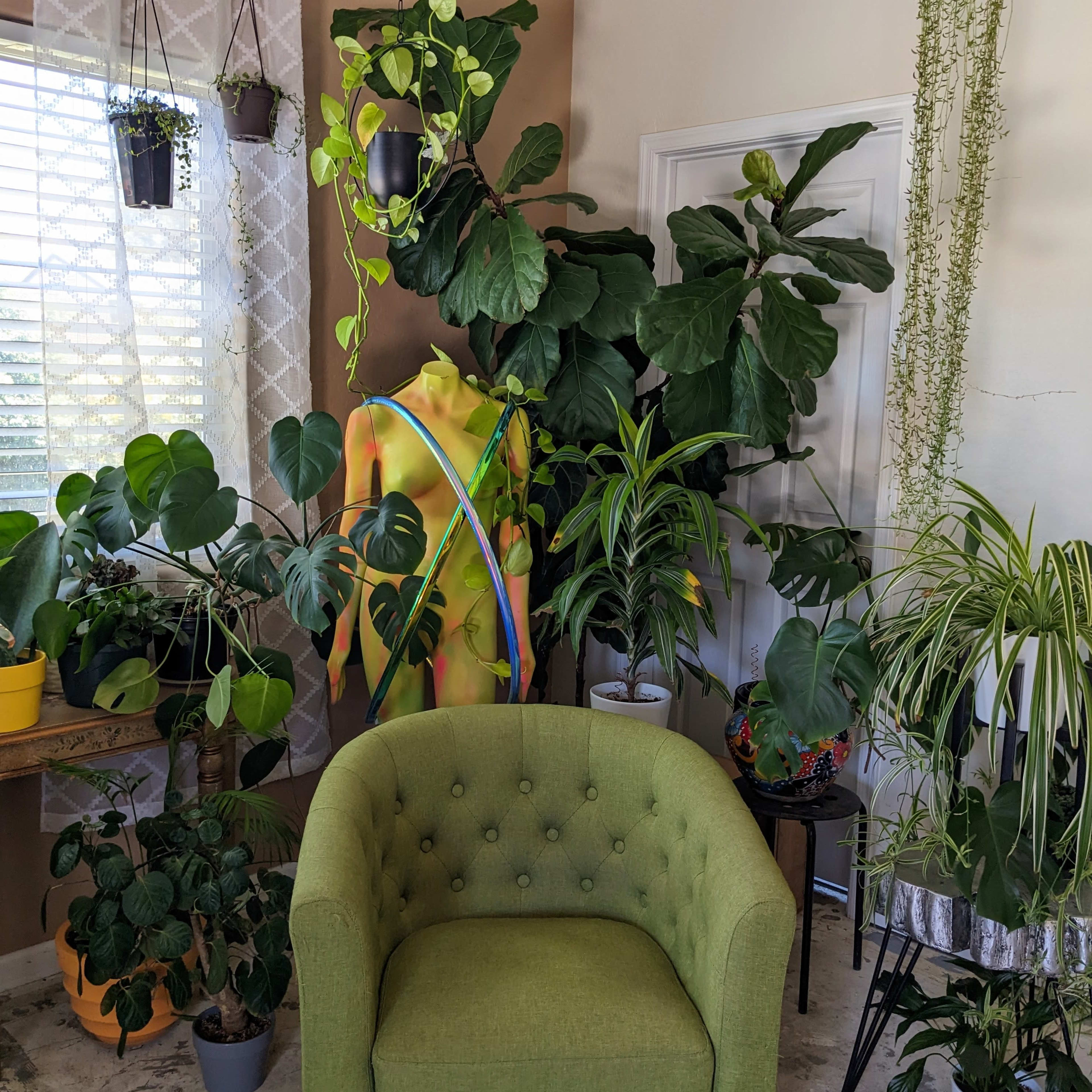 A green upholstered chair is positioned in a room filled with various indoor plants and a colorful mannequin.