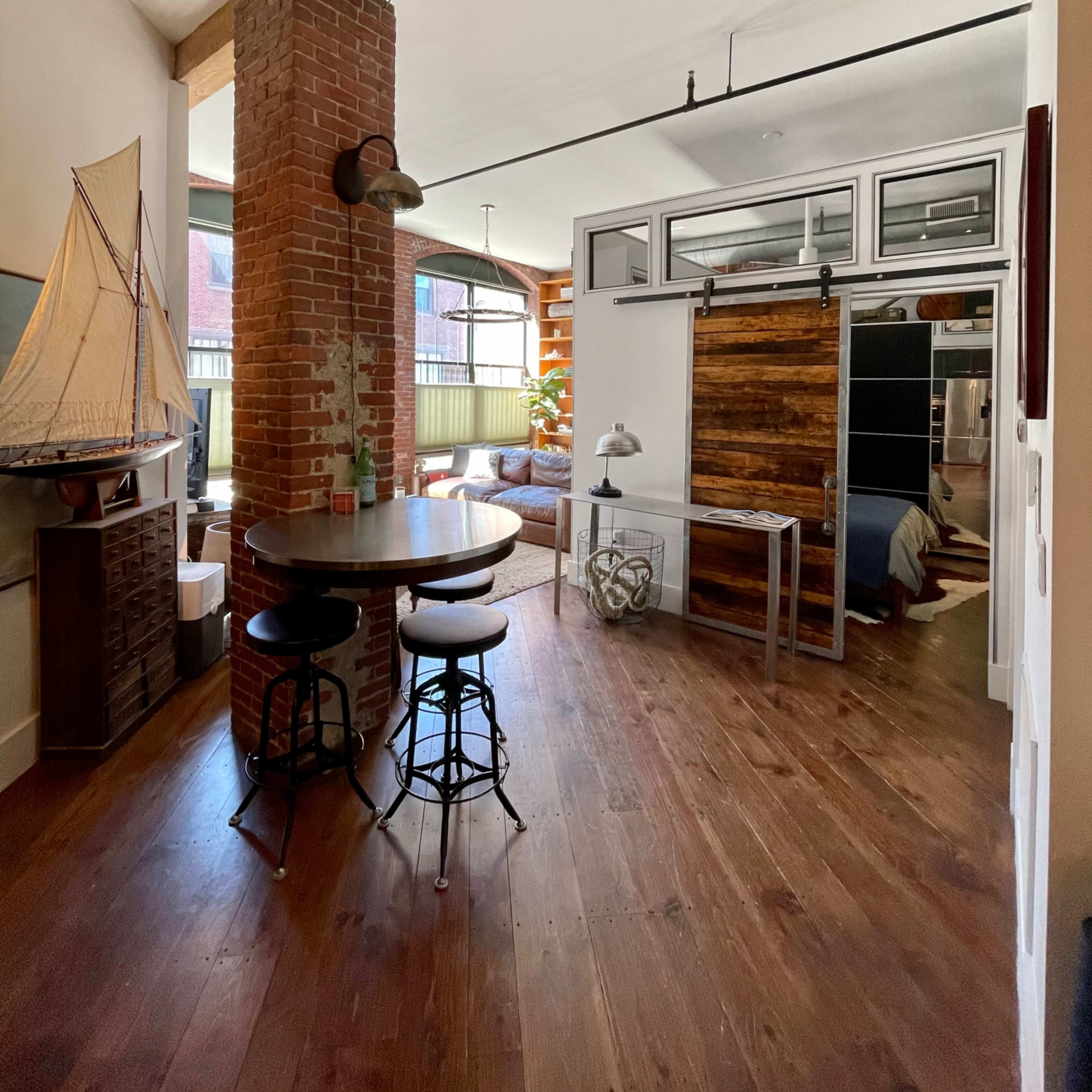 A modern interior space featuring exposed brick walls, wooden beams, and a circular dining table with metal stools, leading to a room with sliding barn doors.
