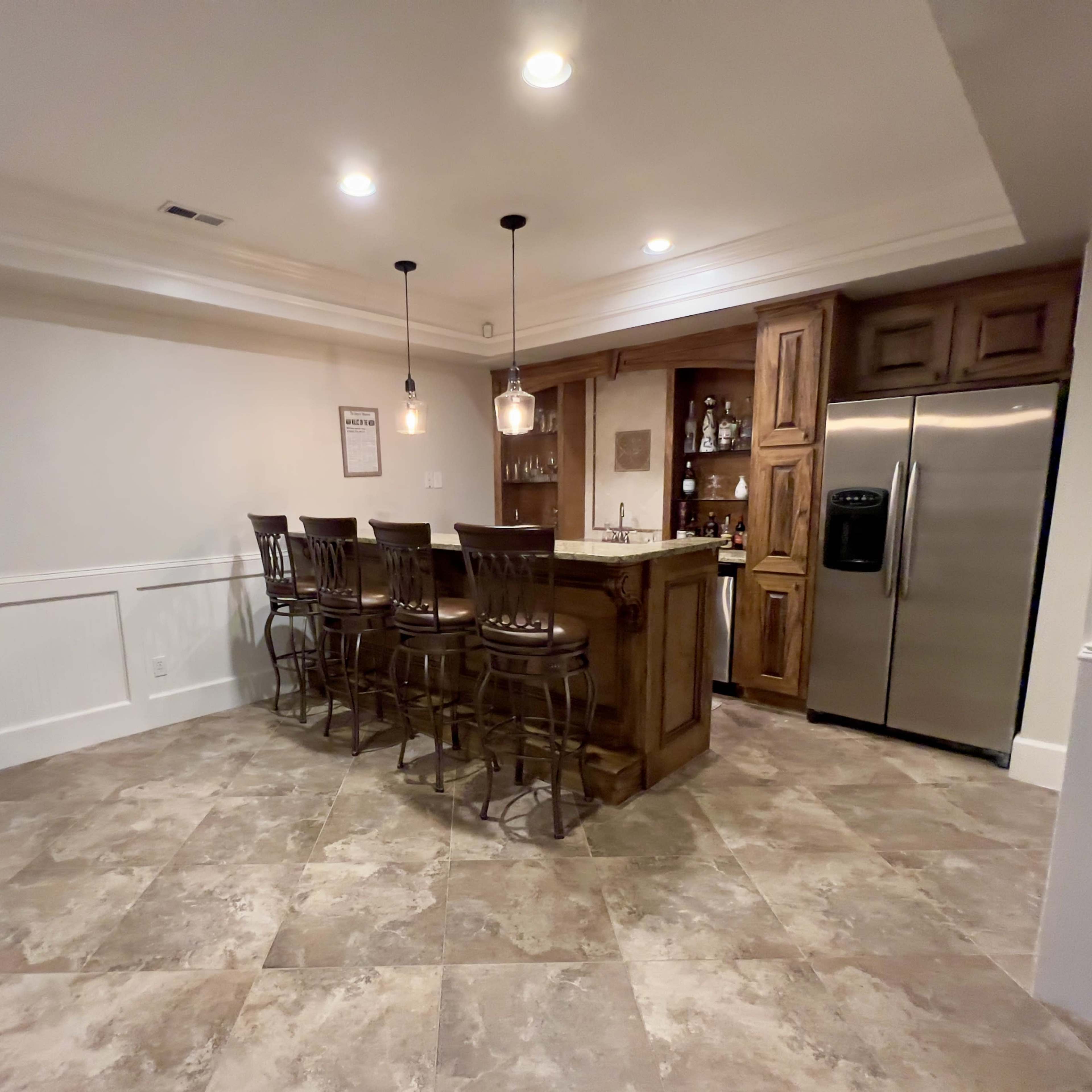 A home bar area with four wooden bar stools, a granite countertop, and shelves displaying various bottles, set against a tiled floor.