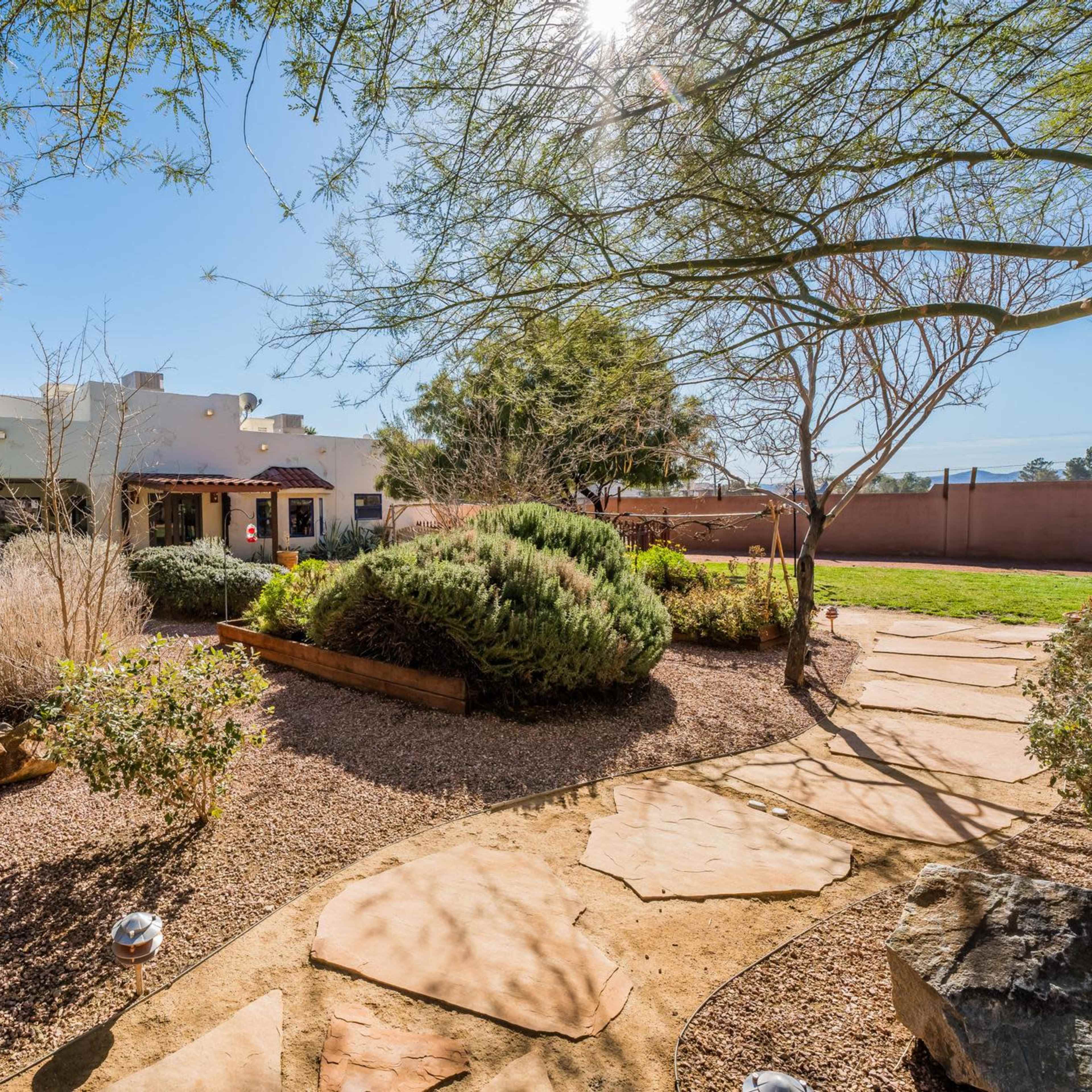 A stone pathway winds through a landscaped yard featuring various shrubs and trees, leading to a house with a southwestern architectural style.