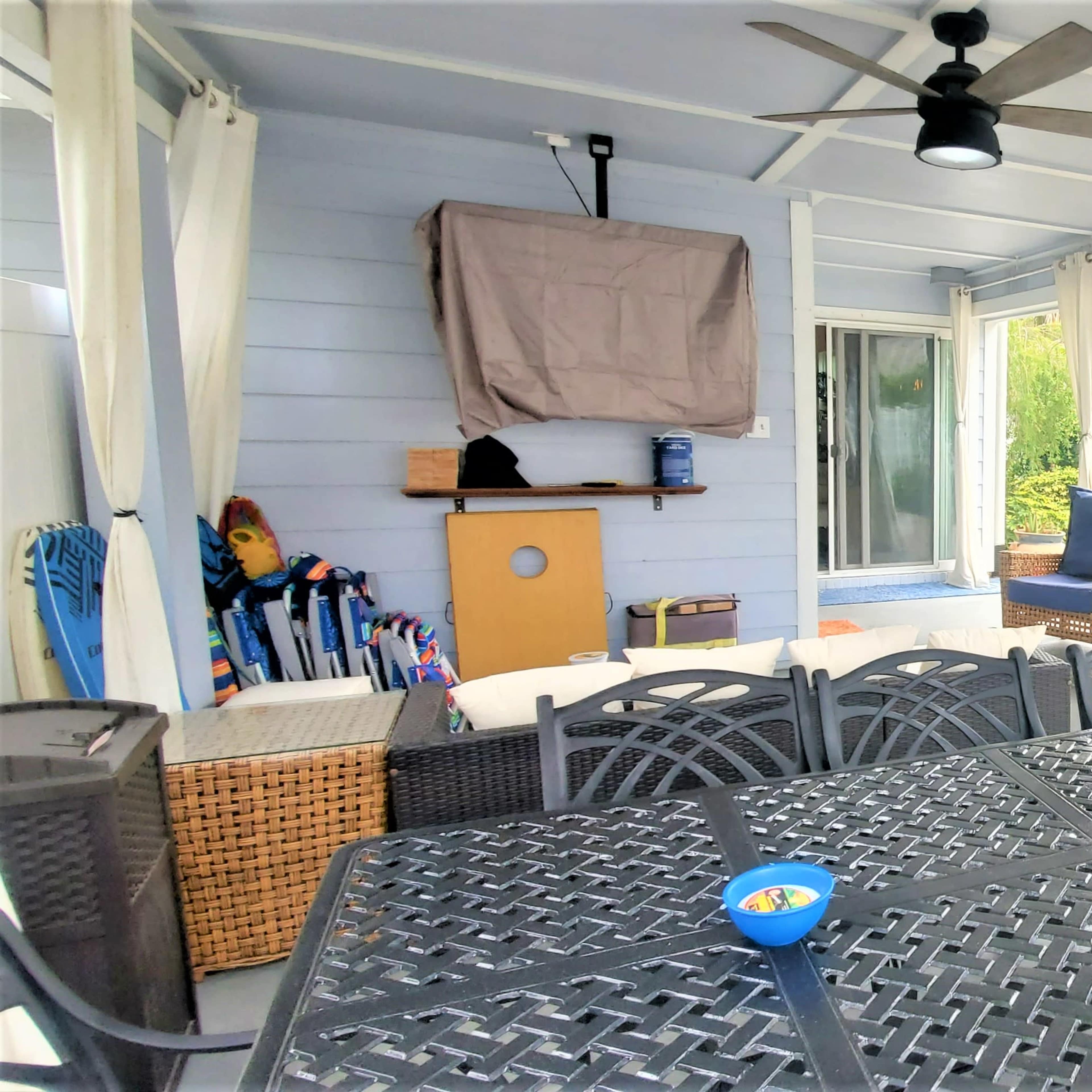 A covered patio area furnished with a large black table, chairs, and a few blue outdoor cushions, surrounded by light-colored curtains and plants.
