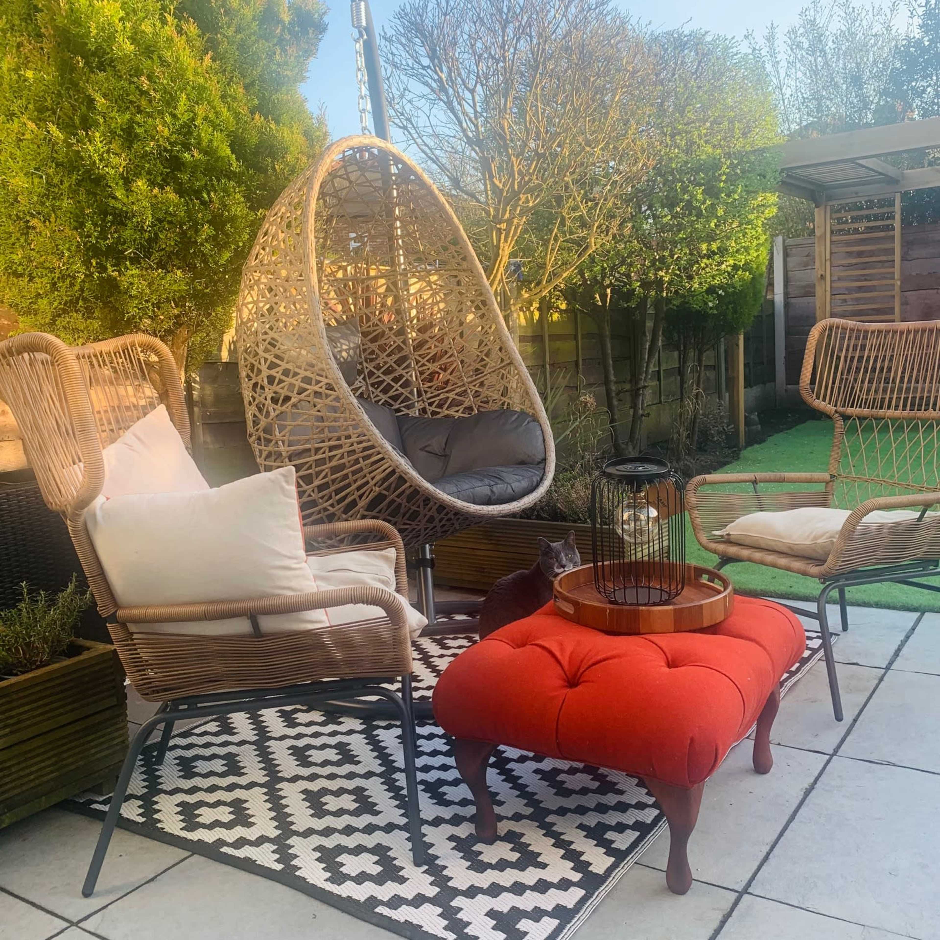 A patio area features a hanging wicker chair, a red upholstered ottoman, and two chairs arranged on a patterned rug surrounded by greenery.