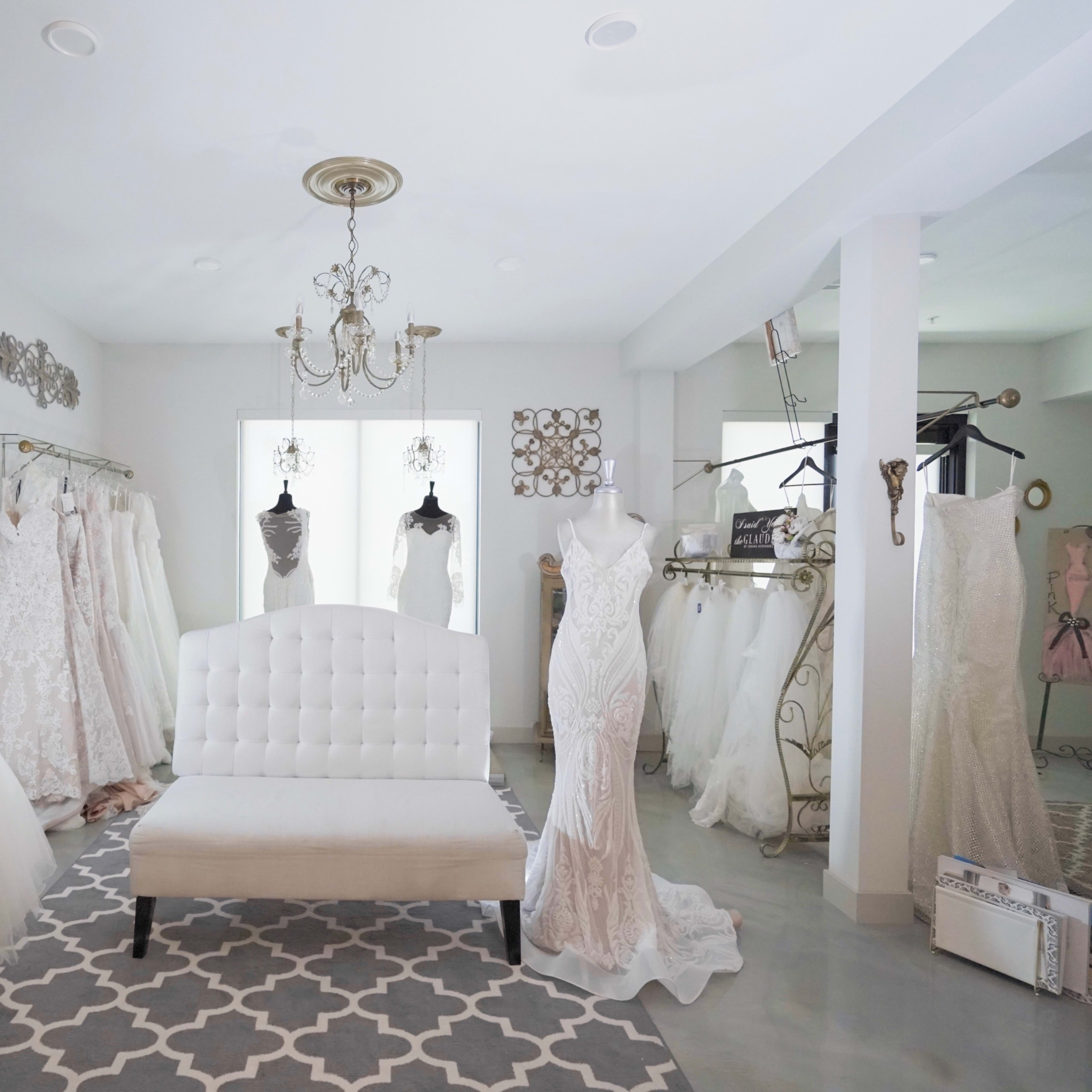 A bridal boutique featuring a variety of wedding dresses displayed on racks, a white seating area in the center, and elegant decorative accents on the walls.