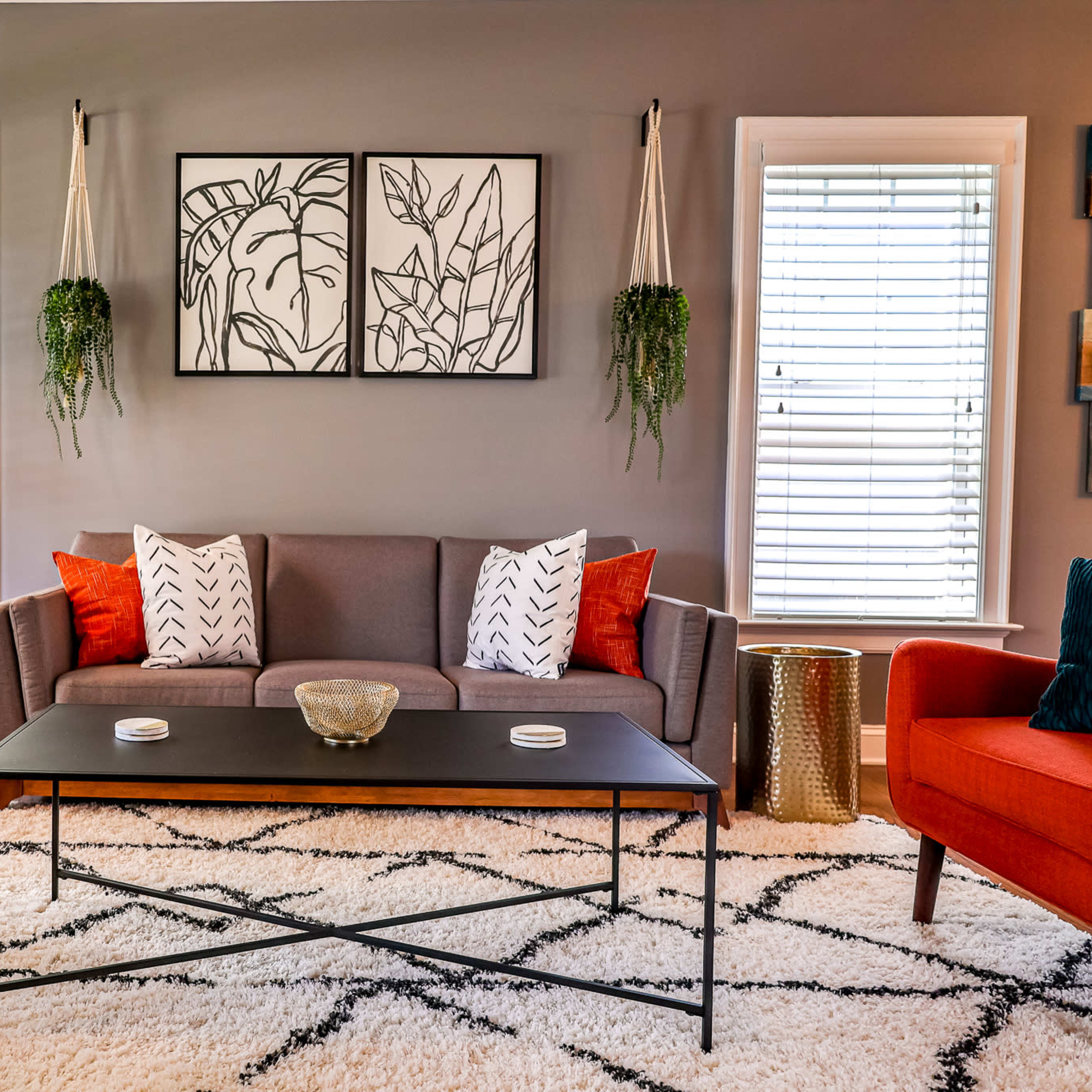 A living room features a gray sofa with colorful cushions, two red accent chairs, a black coffee table, and framed botanical prints on the wall.