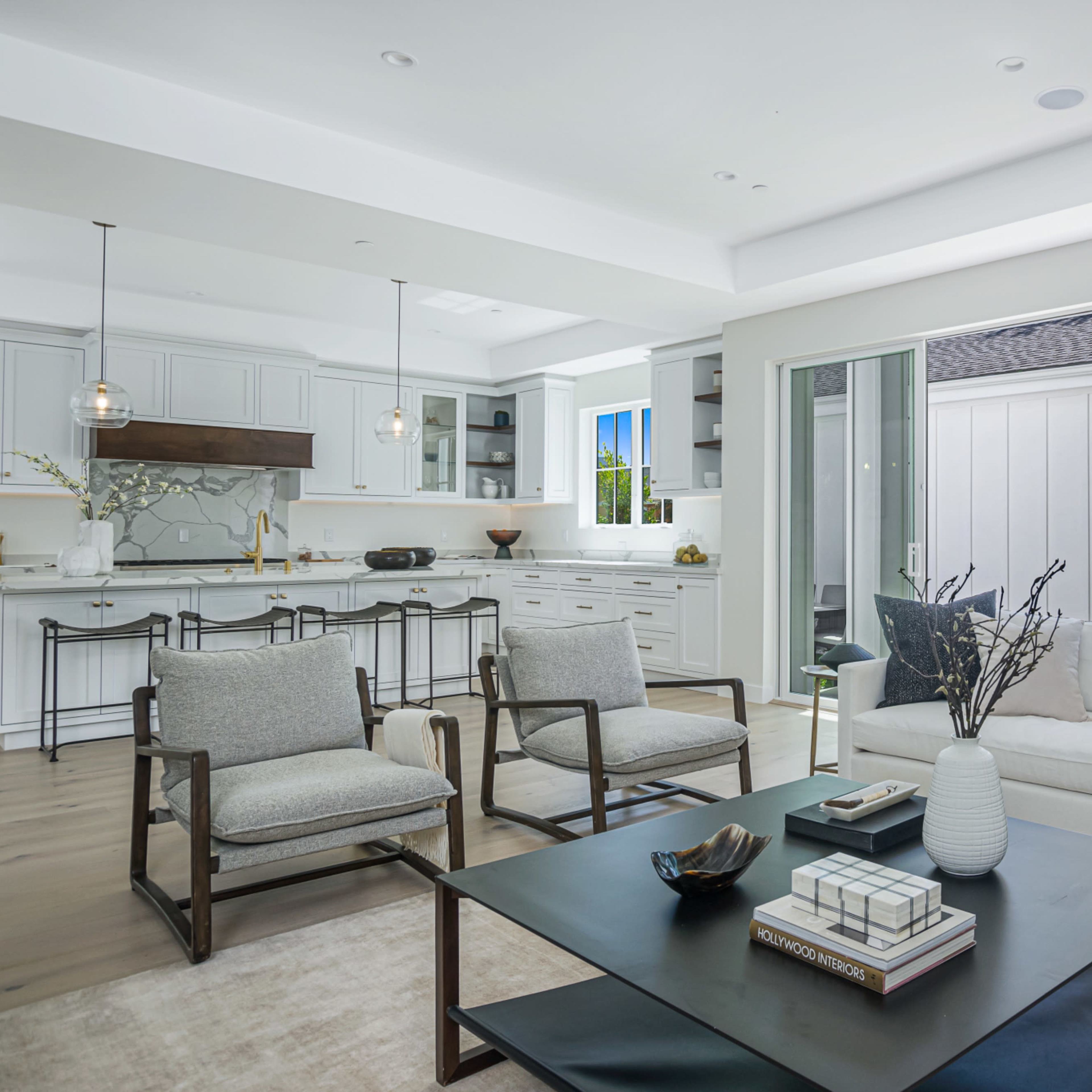 A modern living room features a sectional sofa, two armchairs, and a coffee table, with a view of an adjacent kitchen and outdoor area through large windows.