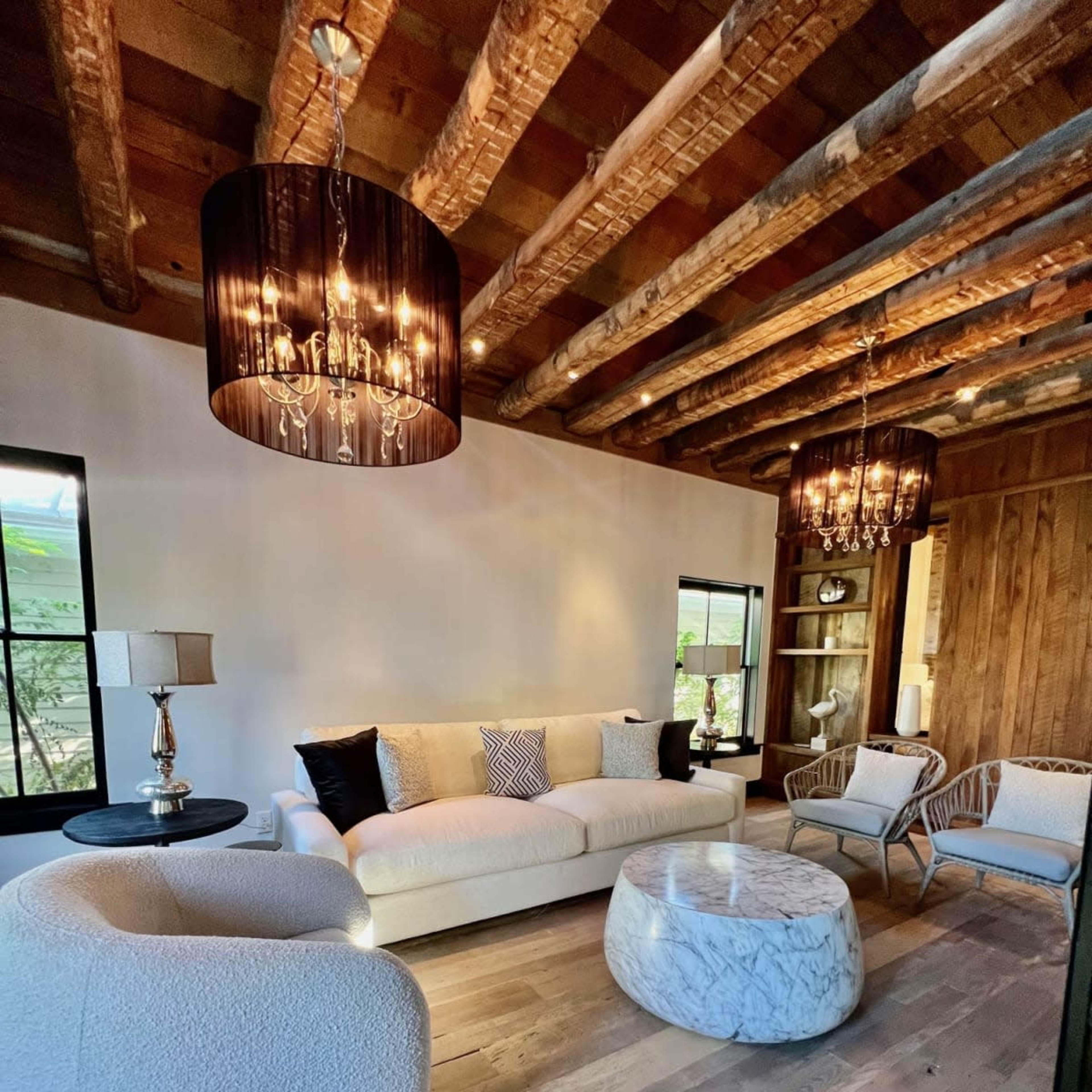 A spacious living room with a large white sofa, two armchairs, a marble coffee table, and wooden ceiling beams, all illuminated by stylish light fixtures.