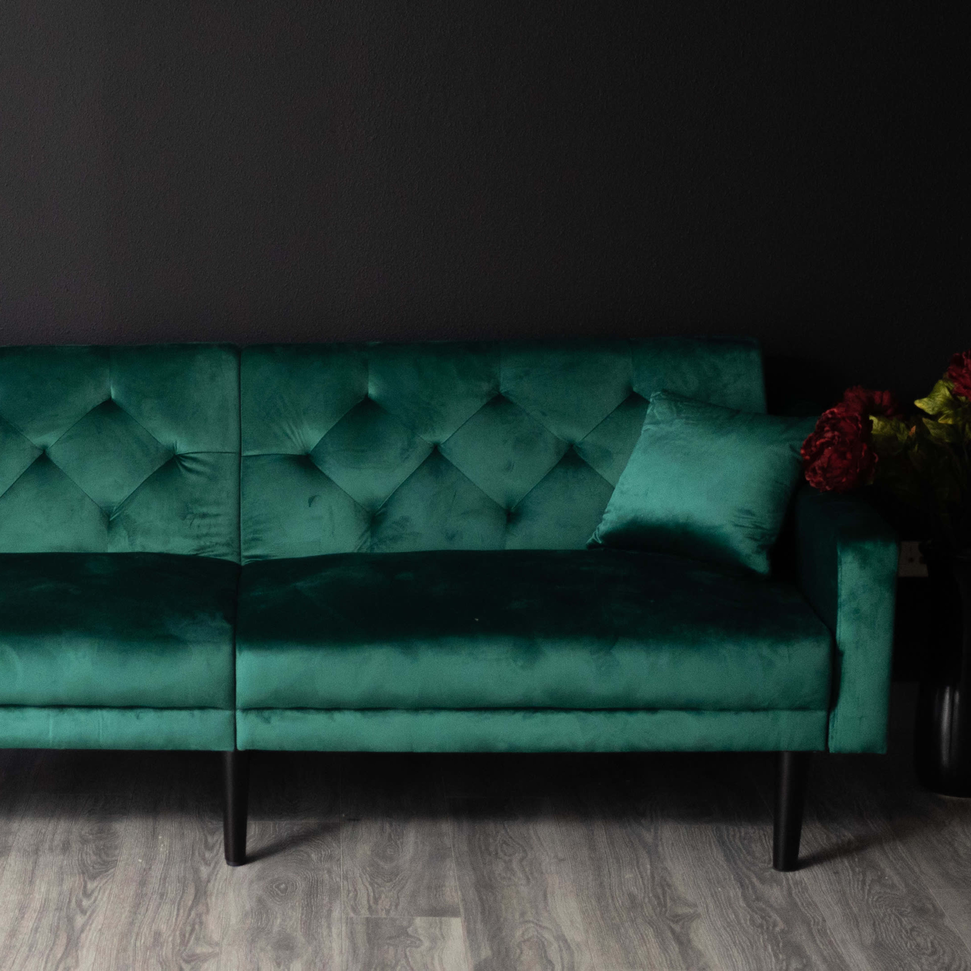 A green velvet sofa with diamond tufting sits against a dark wall, accompanied by a decorative pillow and floral accents.