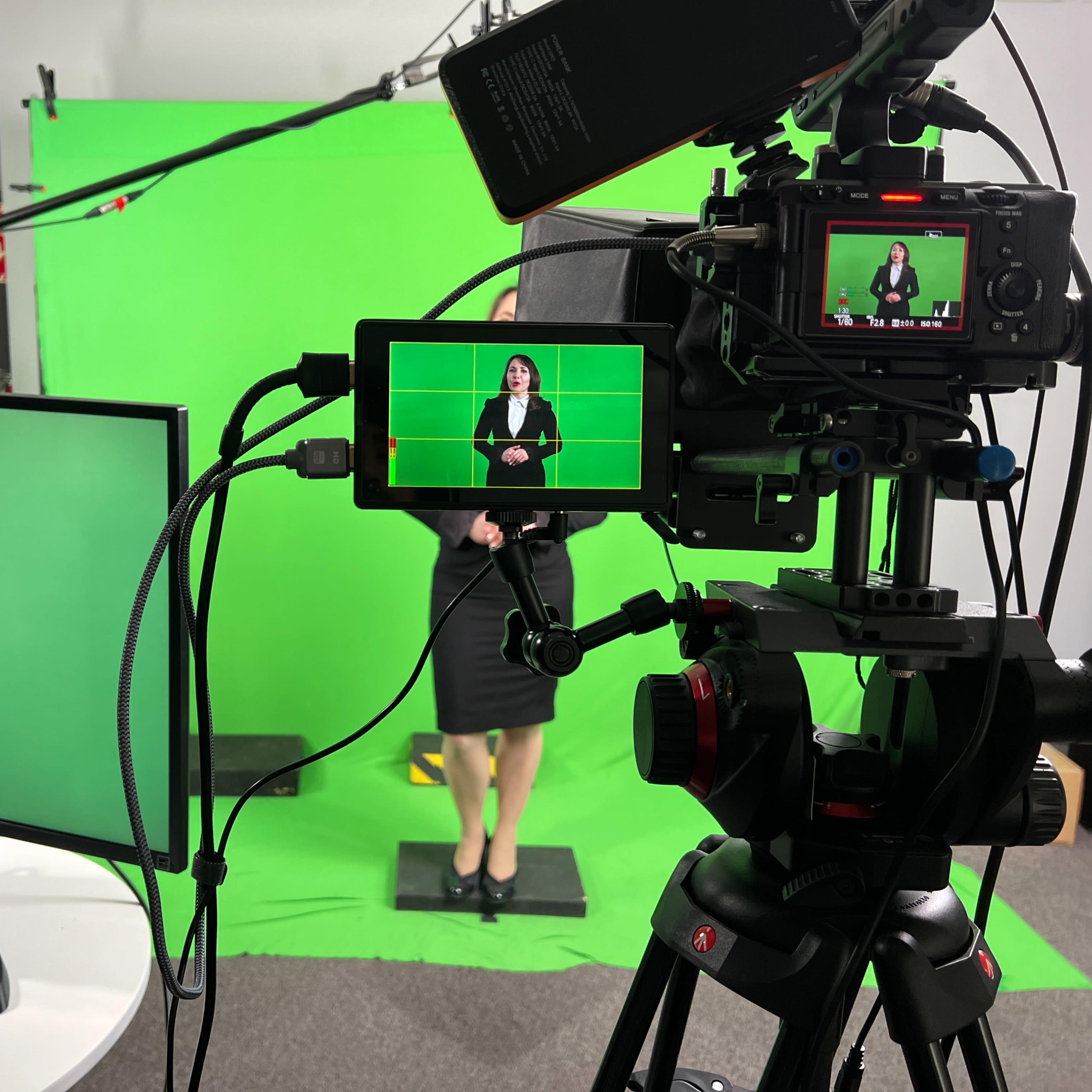 A woman in a black suit stands in front of a green screen while being recorded by cameras on tripods and a monitor displaying her image.